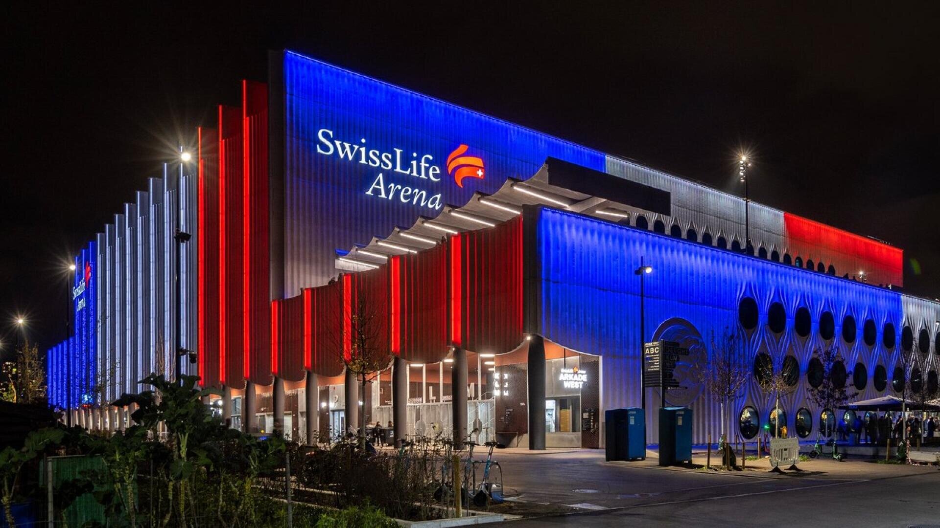 La Swiss Life Arena est illuminée la nuit par des lumières rouges, blanches et bleues sur sa façade ; l'entrée principale et le nom de l'arène sont clairement visibles.