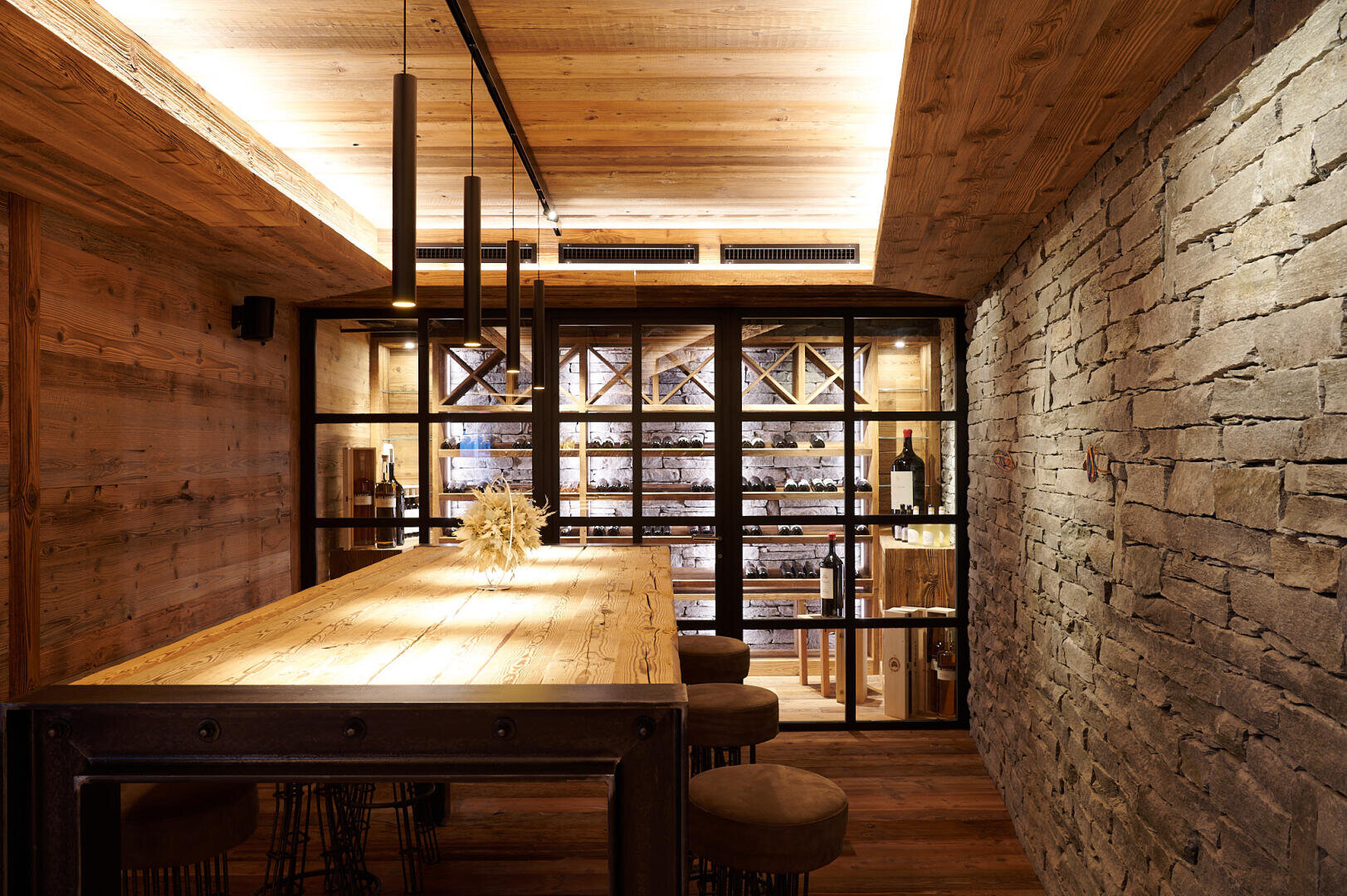 Ein gemütlicher Weinkeller mit Wänden, Decke und Tisch aus Holz, Hängeleuchten im Industrial-Stil, einer Akzentwand aus Stein und Regalen voller Weinflaschen hinter Glastüren. Um den Tisch herum stehen mehrere Hocker.