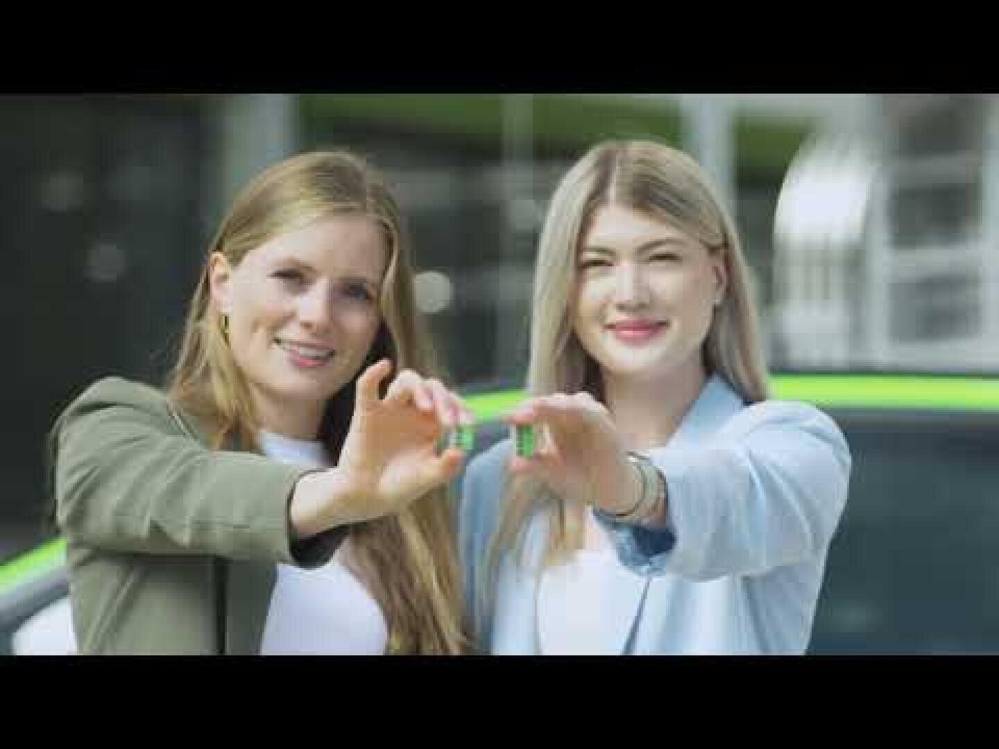 Deux femmes debout à l'extérieur, souriantes, tenant ensemble une petite voiture miniature devant elles, sur un fond flou vert et noir.