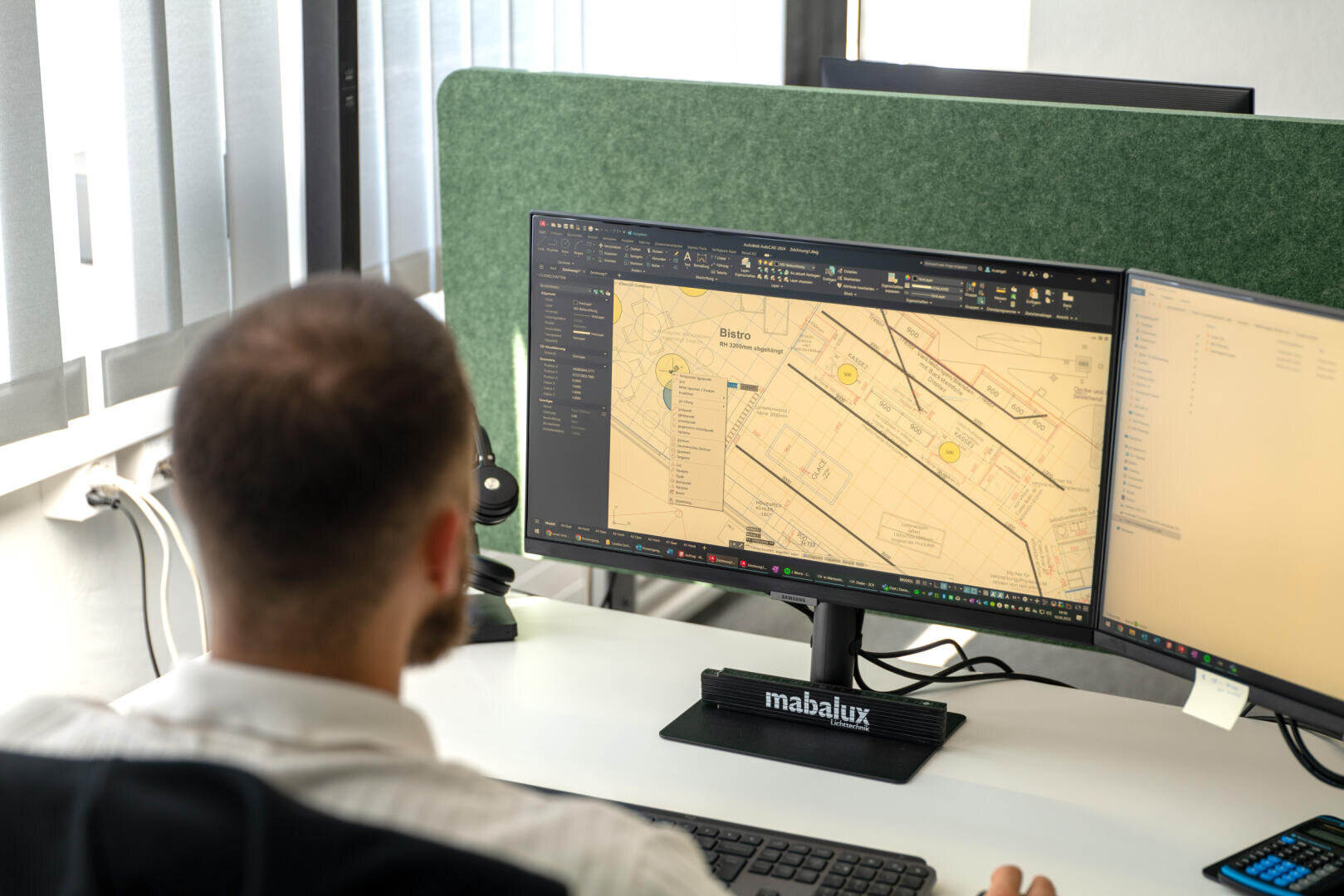 Eine Person sitzt an einem Schreibtisch und arbeitet am Computer, auf dessen großem Monitor eine detaillierte Karte oder ein Architekturplan zu sehen ist. Der Arbeitsplatz ist modern eingerichtet; Trennwände und Büroausstattung sind zu erkennen.