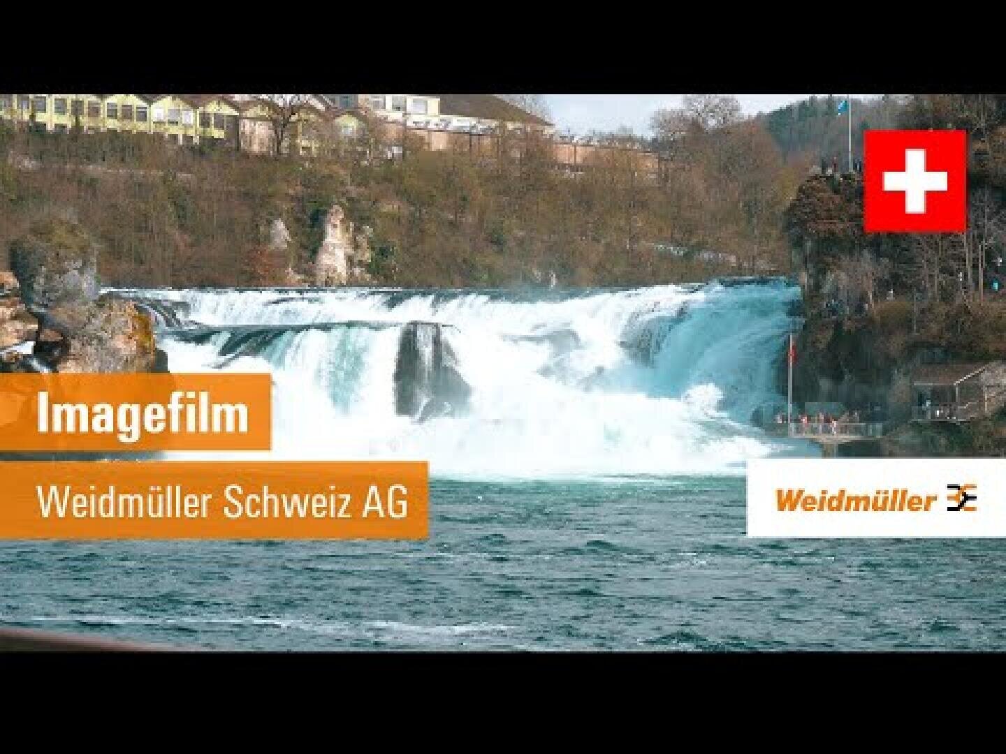 Une large chute d'eau s'écoule sous un ciel nuageux, entourée d'arbres et de bâtiments. En haut à droite, on peut voir le drapeau suisse et le texte orange dit Imagefilm Weidmüller Schweiz AG by Weidmüller.