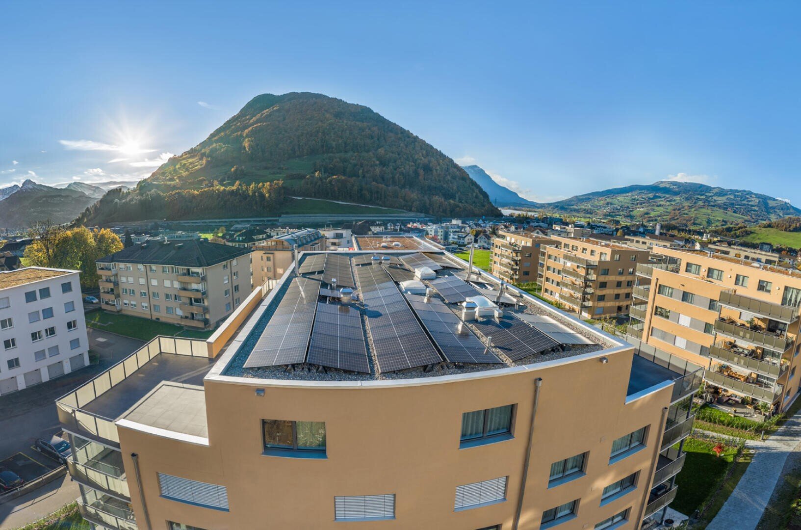 Wohnhäuser mit Solaranlagen auf den Dächern, umgeben von anderen Wohngebäuden, vor dem Hintergrund eines grünen Berges und eines strahlend blauen Himmels an einem sonnigen Tag.