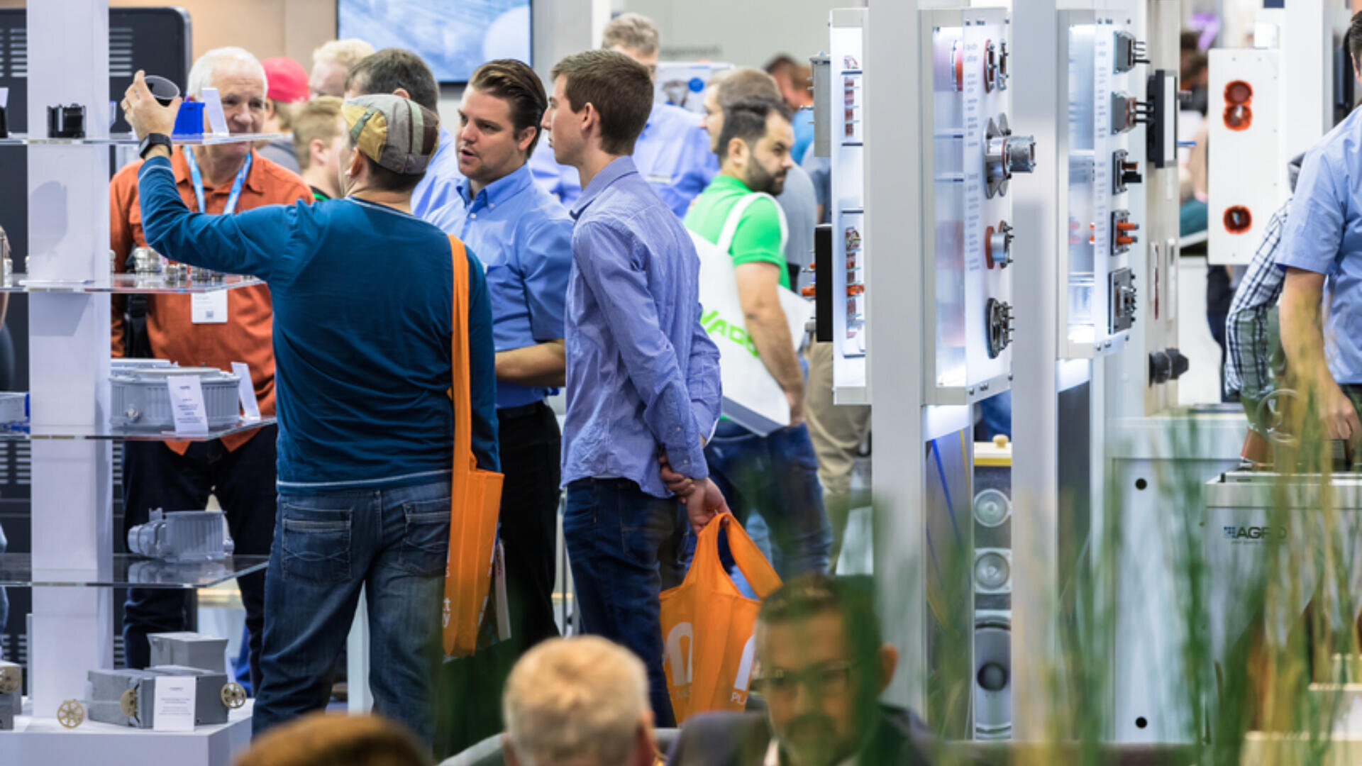 Eine belebte Messehalle mit Menschen, die sich unterhalten, Produkte in Ausstellungsregalen begutachten und orangefarbene Tragetaschen tragen. Im Hintergrund sind Stände und Ausstellungstafeln mit technischen Geräten zu sehen.