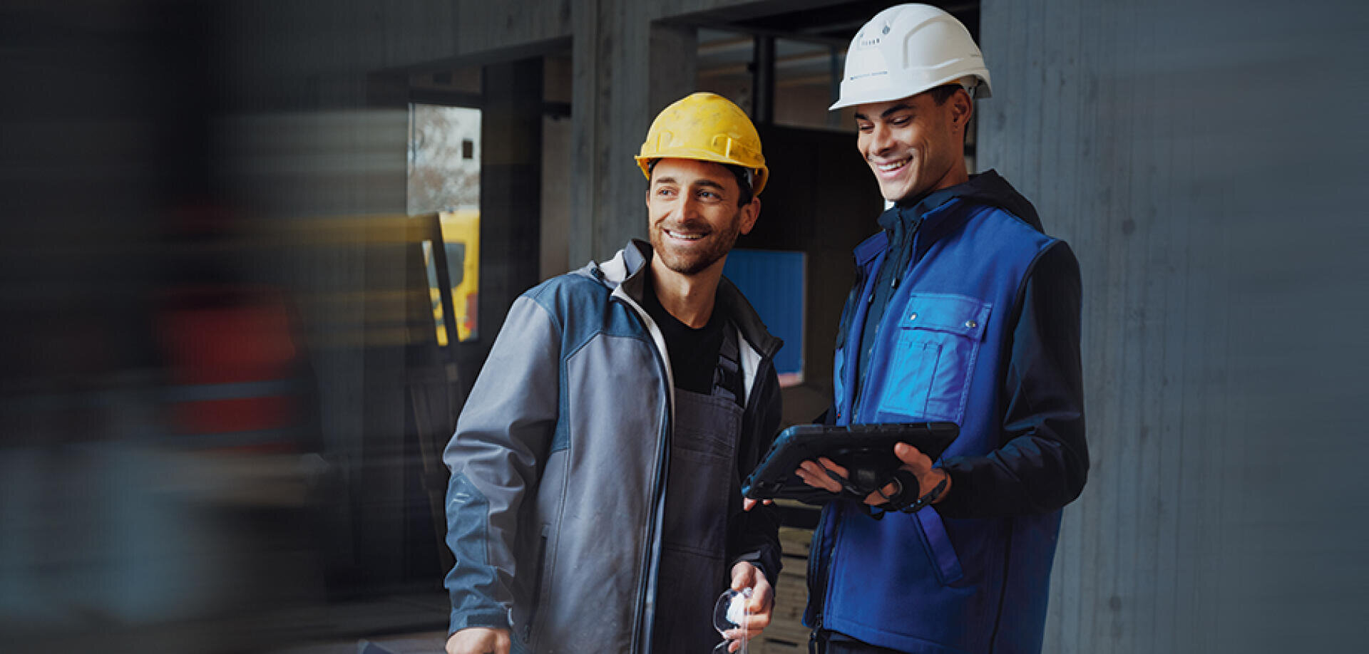 Zwei Bauarbeiter mit Schutzhelmen und Arbeitsjacken stehen in einem Raum und schauen lächelnd auf ein Tablet, was auf die Zusammenarbeit bei einem Bauprojekt hindeutet.