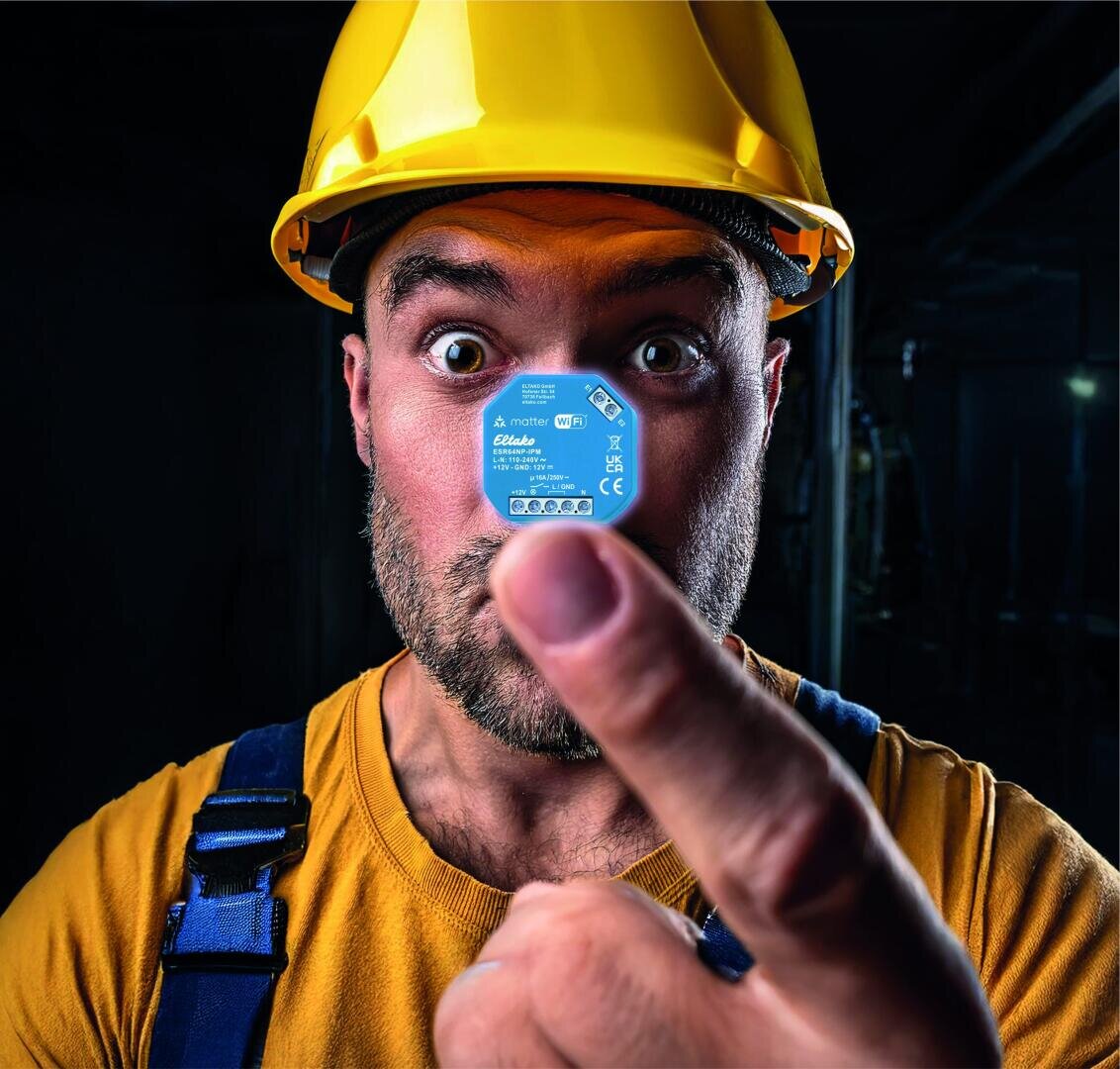 Ein Bauarbeiter mit gelbem Schutzhelm und gelbem Hemd hält ein kleines blaues Elektronikbauteil dicht vor sein Gesicht und betrachtet es mit weit aufgerissenen Augen. Der Hintergrund ist dunkel und unscharf.