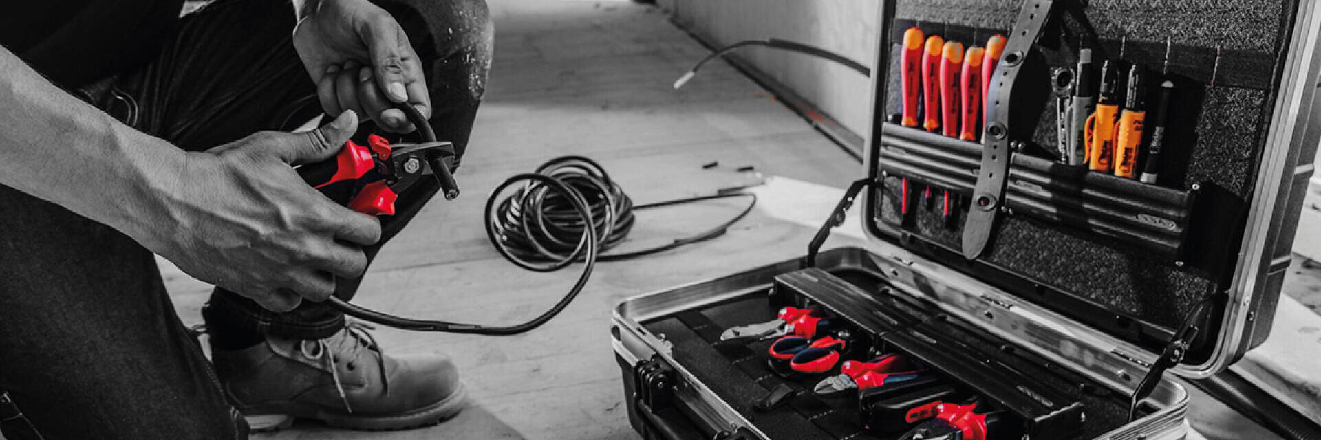 Une personne utilise un coupe-fil rouge et noir sur un câble près d'une boîte à outils ouverte remplie de différents outils à main ; l'image met en évidence les outils en couleur, tandis que le reste est en noir et blanc.
