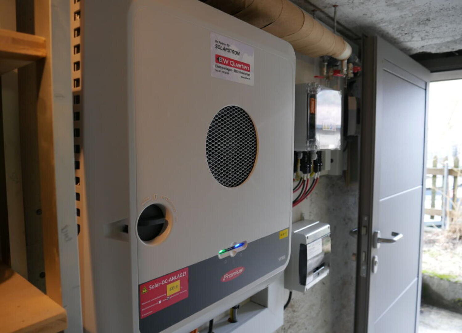 Eine Nahaufnahme eines Solarwechselrichters und elektrischer Schalttafeln, die an einer Wand in einem Hauswirtschaftsraum mit offener Tür montiert sind, die Tageslicht hereinlässt.