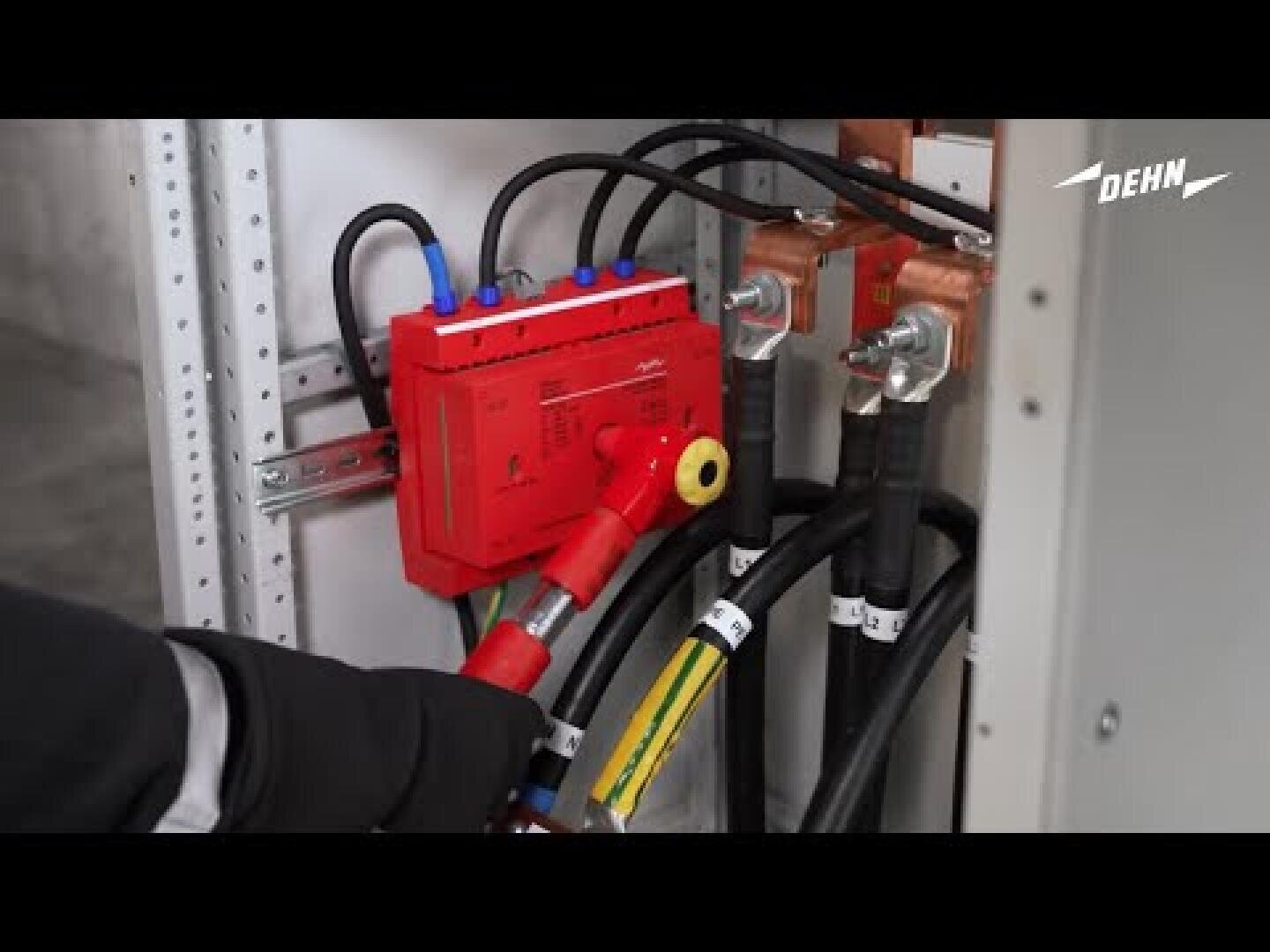 Eine behandschuhte Hand hält ein rotes elektrisches Prüfgerät, das an dicke schwarze Kabel in einer Schalttafel angeschlossen ist. In der rechten oberen Ecke ist die Marke DEHN zu sehen.