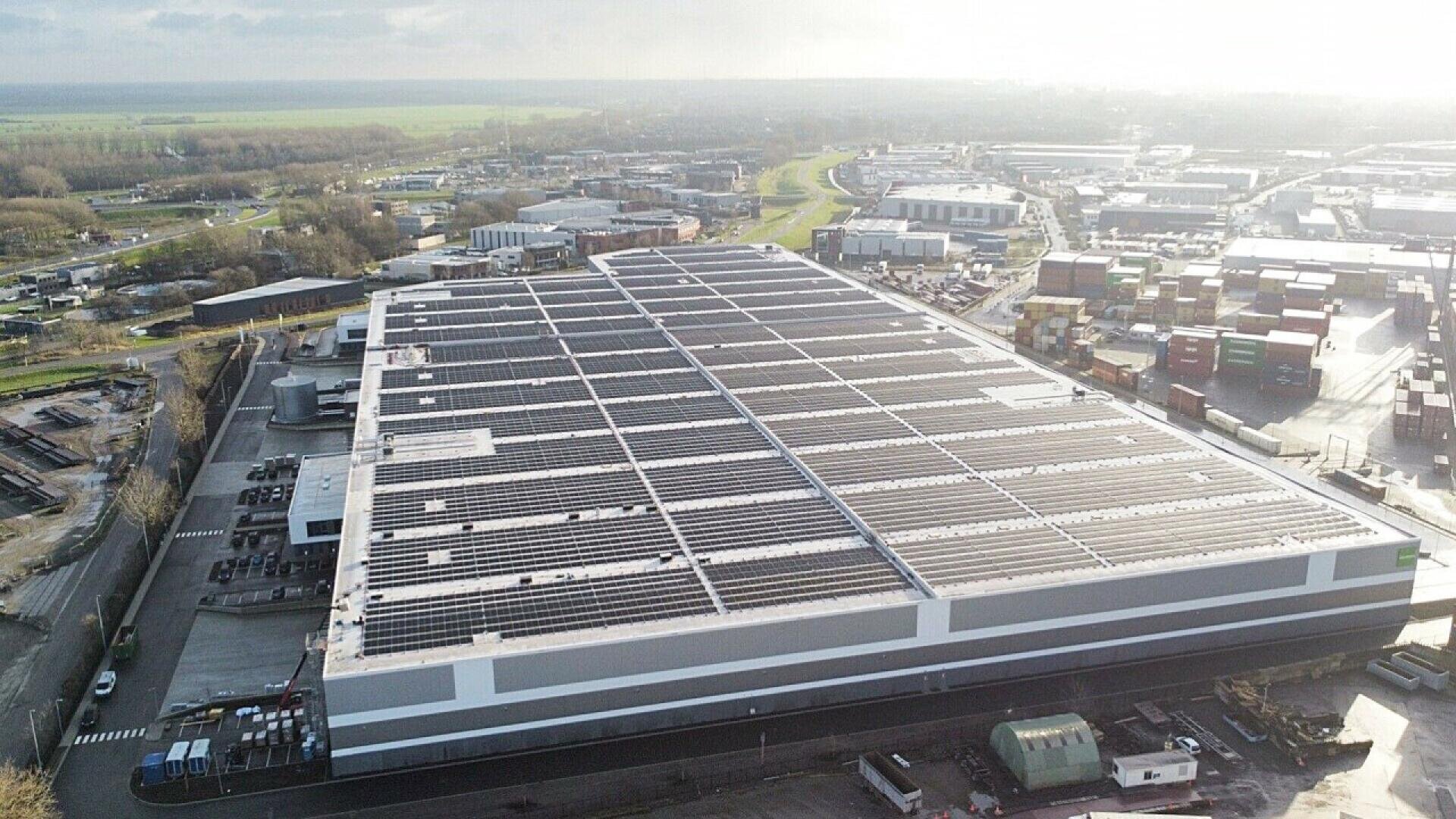 Un grand entrepôt industriel dont le toit est recouvert de panneaux solaires, entouré de routes, de véhicules et d'autres bâtiments dans une zone industrielle, vu d'une perspective aérienne par une journée ensoleillée.