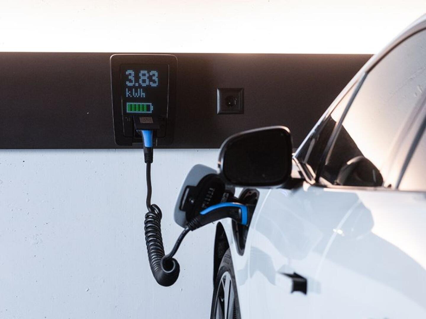 Une voiture électrique blanche est en train de se recharger sur une station de recharge murale qui affiche 3,83 kWh. Le câble de recharge est branché sur la voiture et l'indicateur de batterie indique une charge partielle.