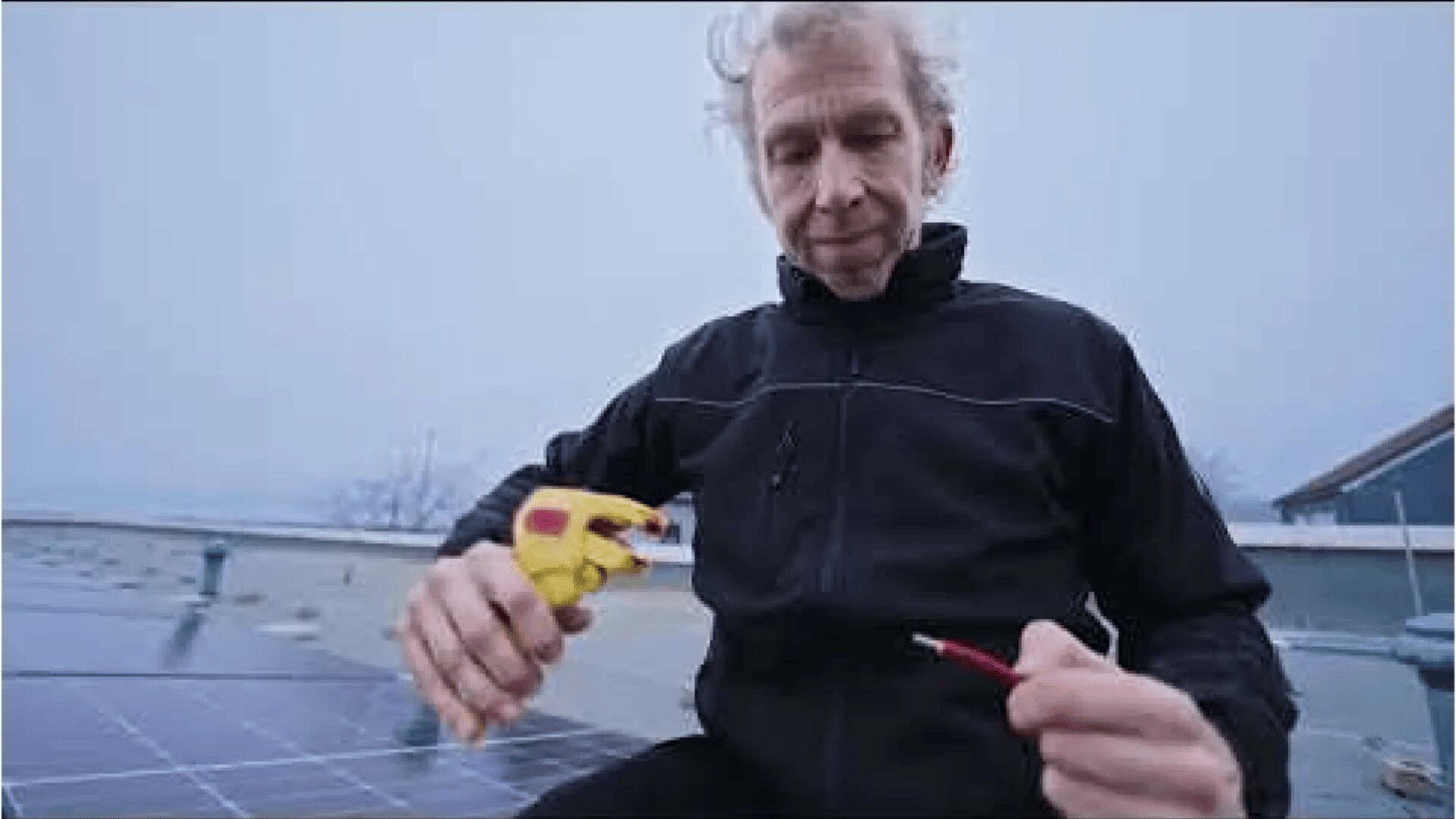 Une personne vêtue d'une veste noire est assise sur un toit à côté de panneaux solaires, tenant un flacon pulvérisateur dans une main et un fil rouge dans l'autre, semblant effectuer des travaux d'entretien ou de nettoyage.