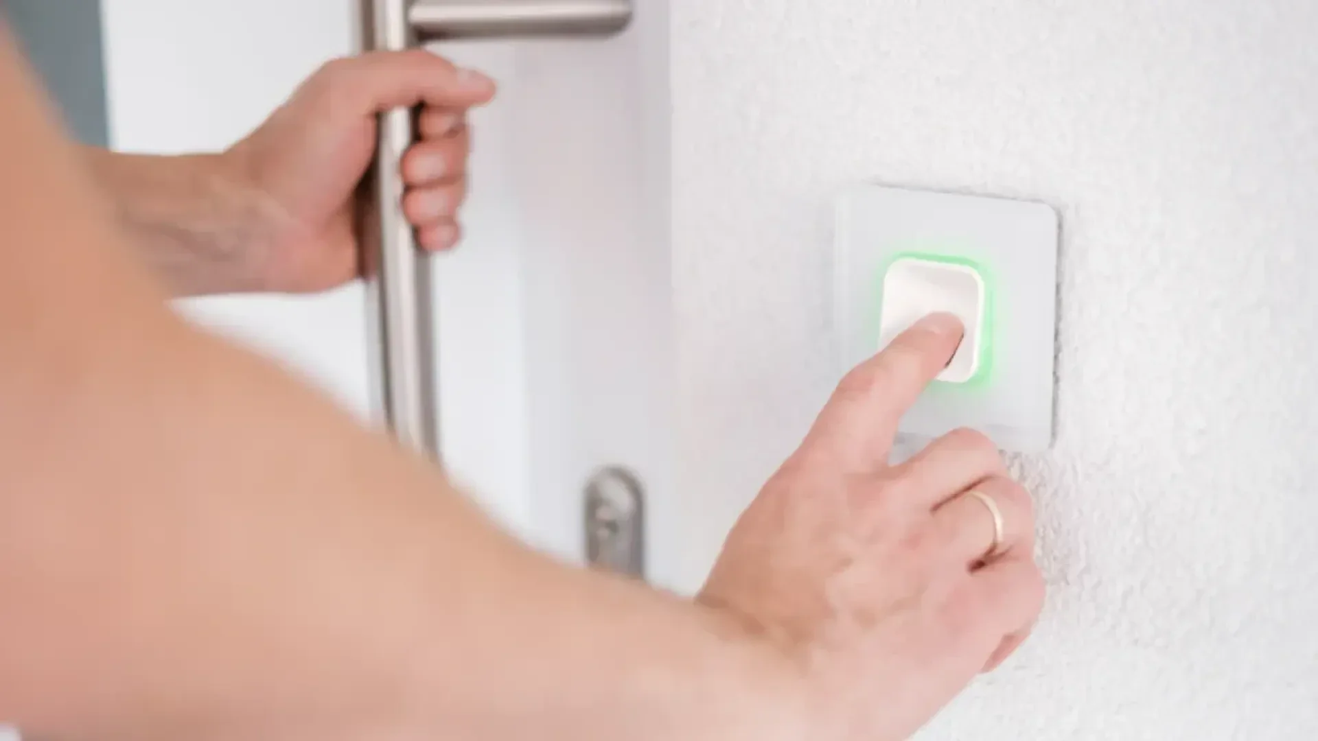 Une personne appuie d'une main sur une sonnette ou un interrupteur allumé en vert sur un mur blanc, tout en tenant la poignée de la porte de l'autre main.