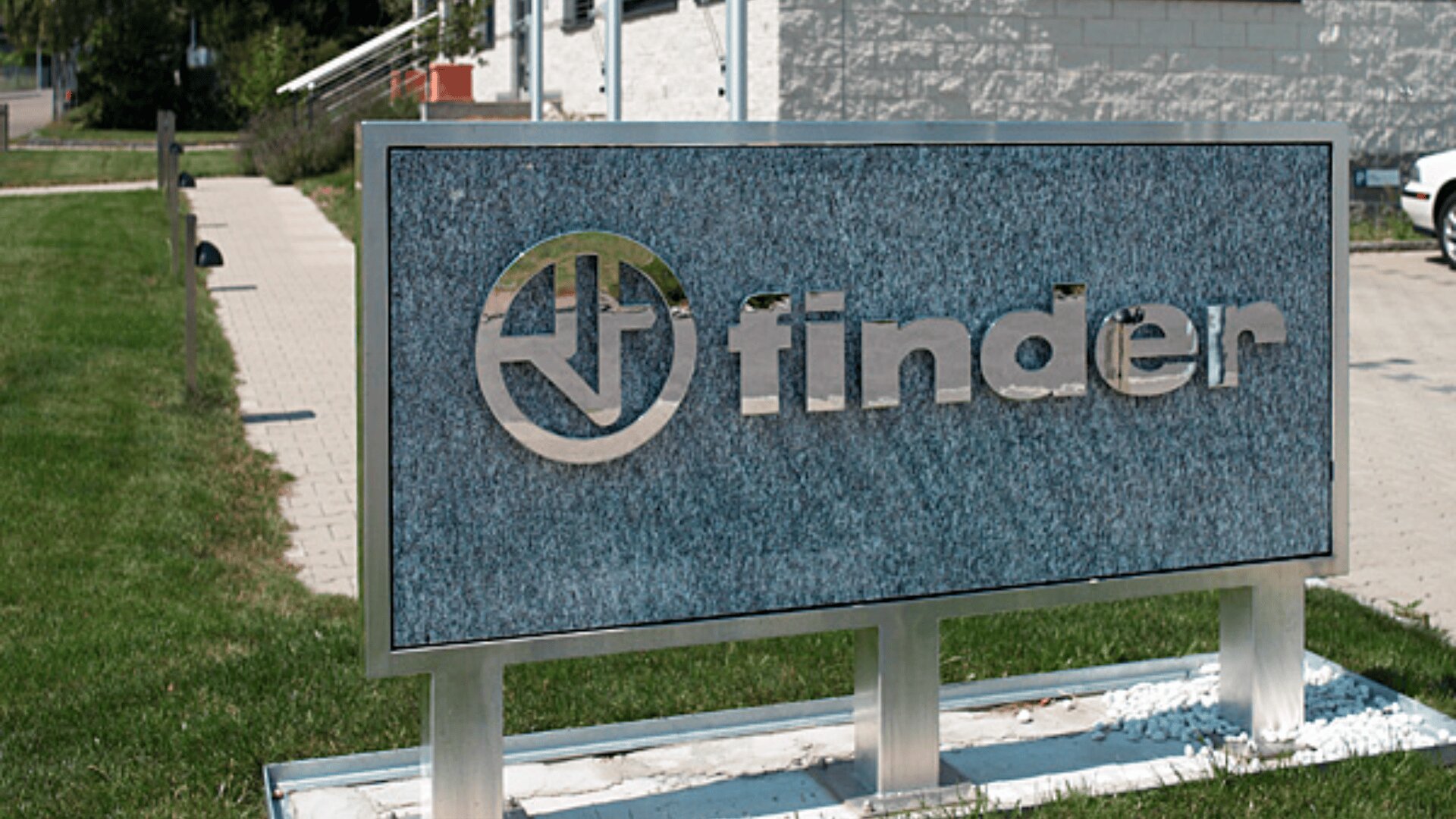 Ein steinernes Schild mit dem Wort "Finder" und einem kreisförmigen Logo steht vor einem Gebäude, umgeben von Gras und einem Gehweg.