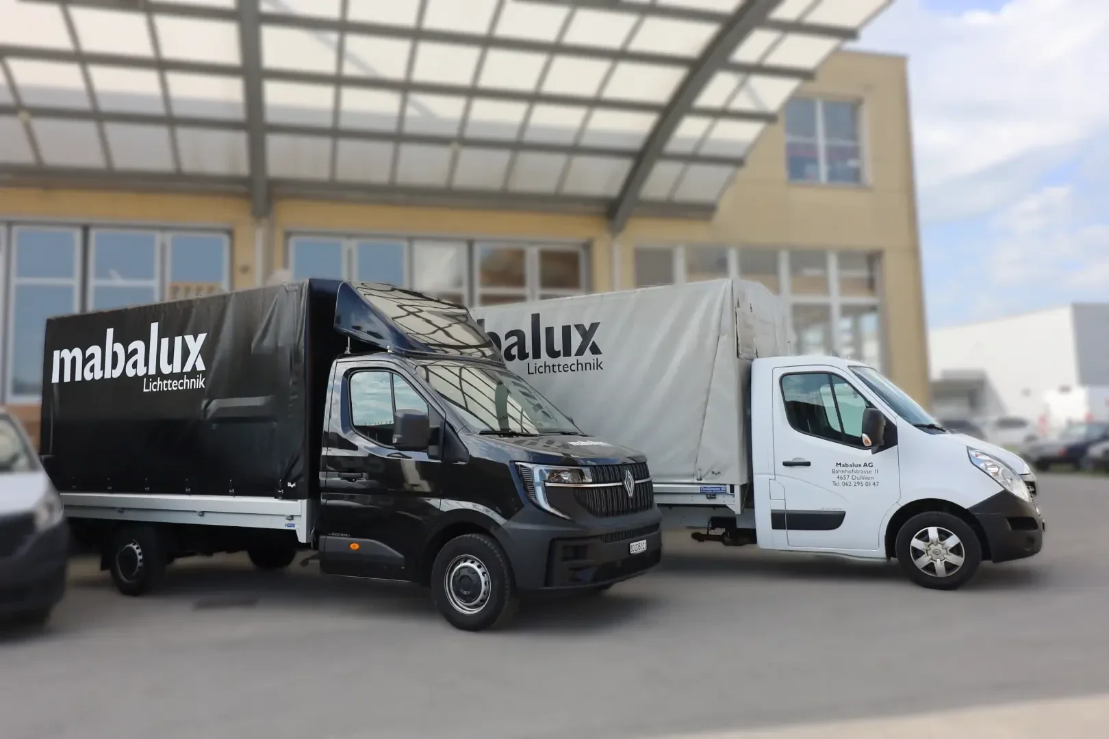 Vor einem Industriegebäude mit großen Fenstern und einem Glasvordach stehen zwei Lieferwagen mit der Aufschrift „mabalux Lichttechnik“ nebeneinander.