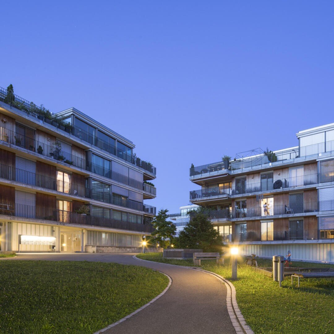Moderne Wohnblocks mit Balkonen bei Einbruch der Dämmerung, umgeben von grünen Rasenflächen und einem geschwungenen Weg im Vordergrund. Einzelne LIchter am Weg und in den Häusern leuchten.