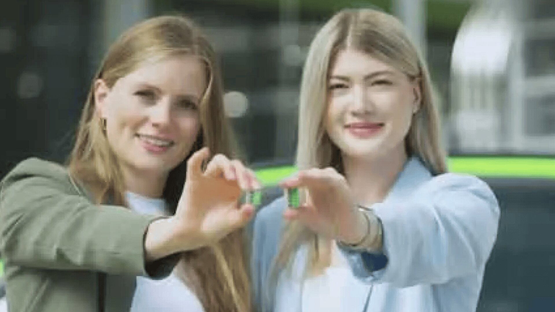 Zwei Frauen, die lächelnd einen kleinen grünen Gegenstand in der Hand halten, stehen im Freien vor einem verschwommenen Hintergrund, hinter dem ein grünes Fahrzeug zu sehen ist.