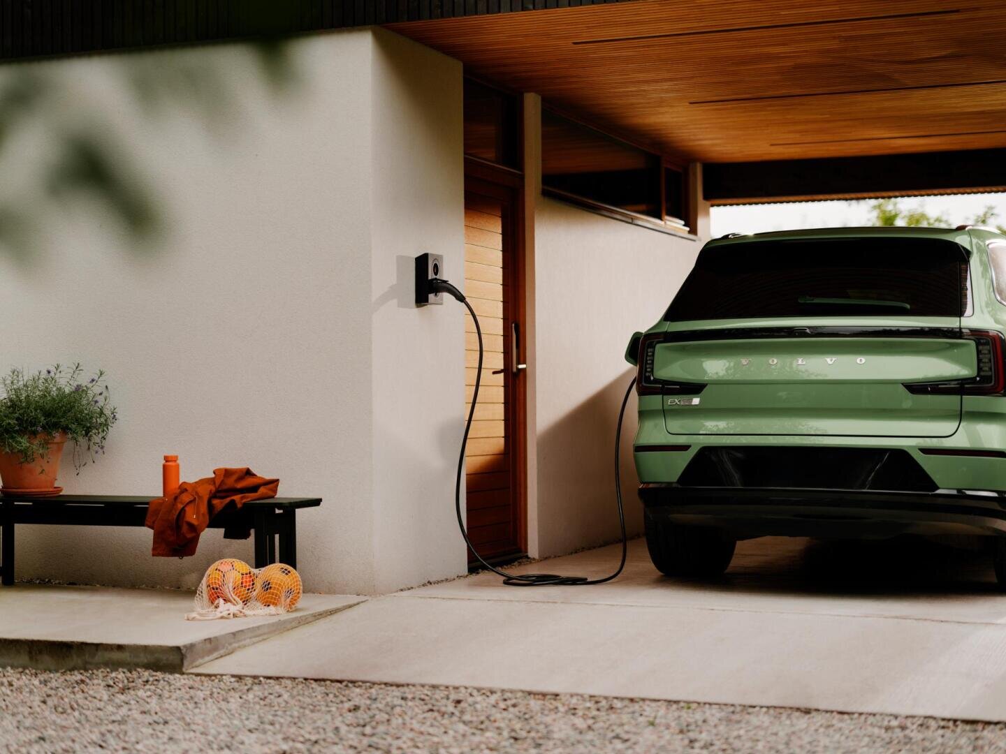 Un SUV électrique Volvo de couleur verte est garé sous un abri moderne et branché à une borne de recharge murale. À côté se trouve un banc sur lequel sont posés une plante en pot, une bouteille d'eau orange et des équipements de sport.