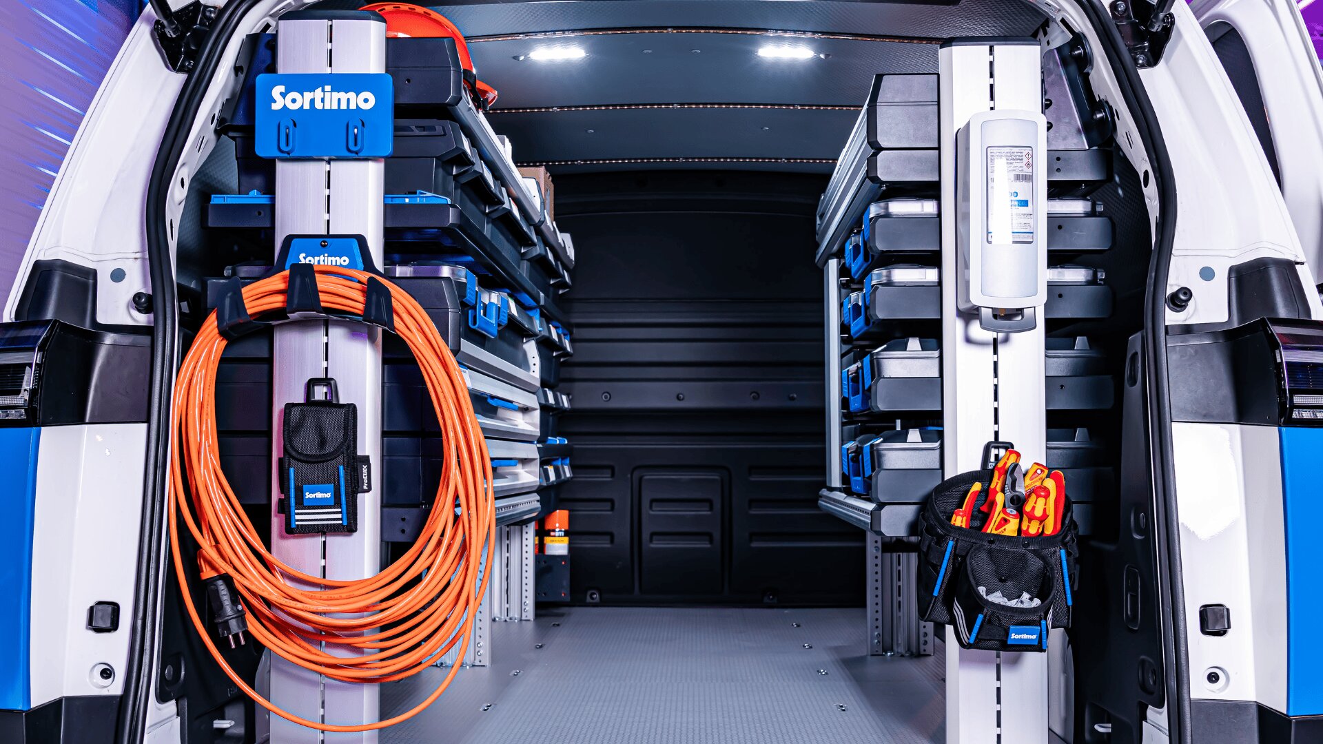 Das Bild zeigt das geordnete Innere eines Arbeitswagens mit Regalen mit blauen Ablagefächern, orangefarbenen Verlängerungskabeln, Werkzeugen und Sicherheitsausrüstung, die an beiden Seiten angebracht sind. Der Lieferwagen ist sauber und gut beleuchtet, was auf eine professionelle Einrichtung schließen lässt.