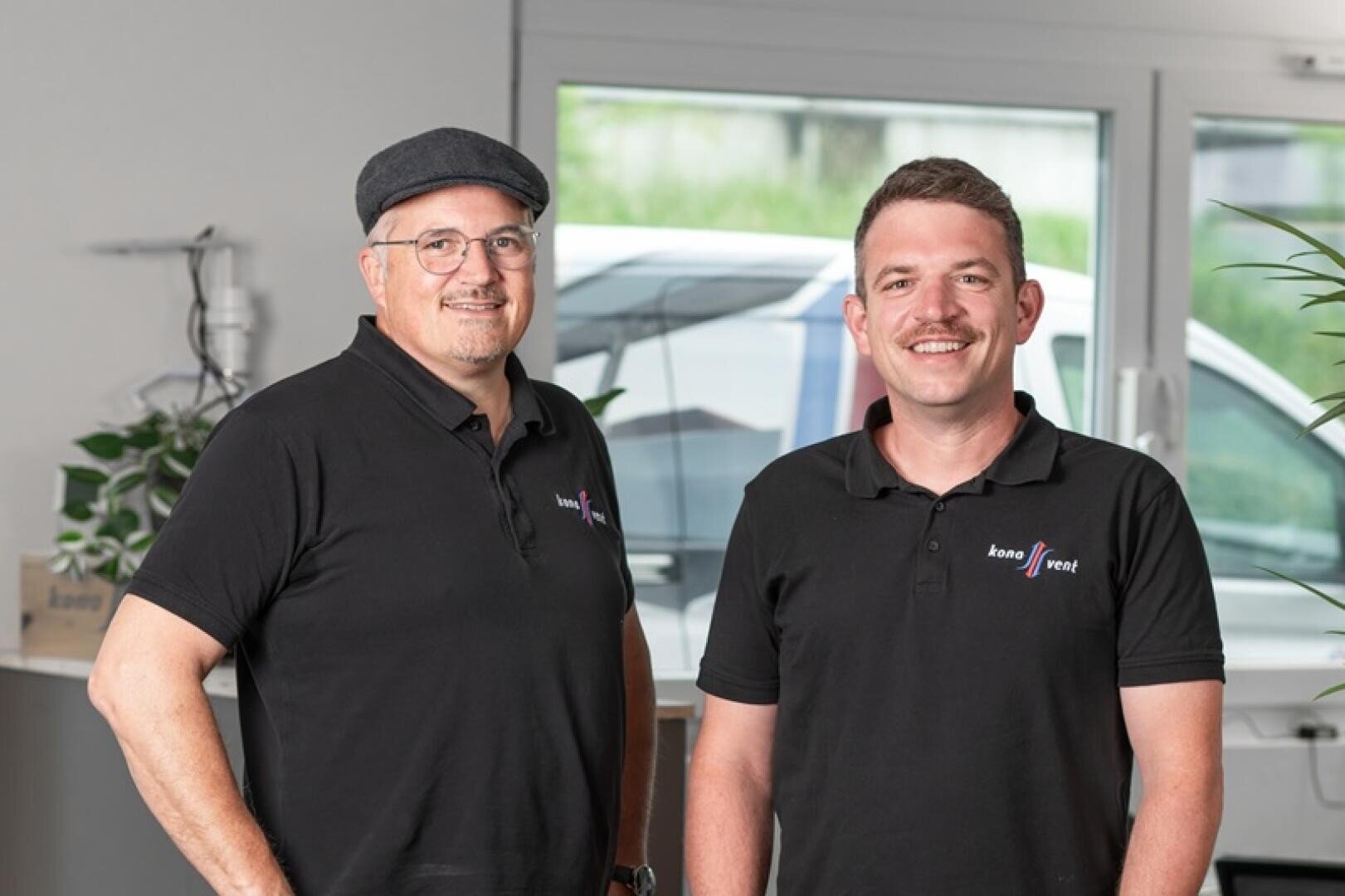 Zwei Männer in schwarzen Polohemden mit aufgestickten Logos stehen lächelnd in einem Innenraum in der Nähe eines Fensters, einer trägt eine flache Kappe und eine Brille, der andere einen Schnurrbart. Im Hintergrund sind Pflanzen und ein geparktes Fahrzeug zu sehen.