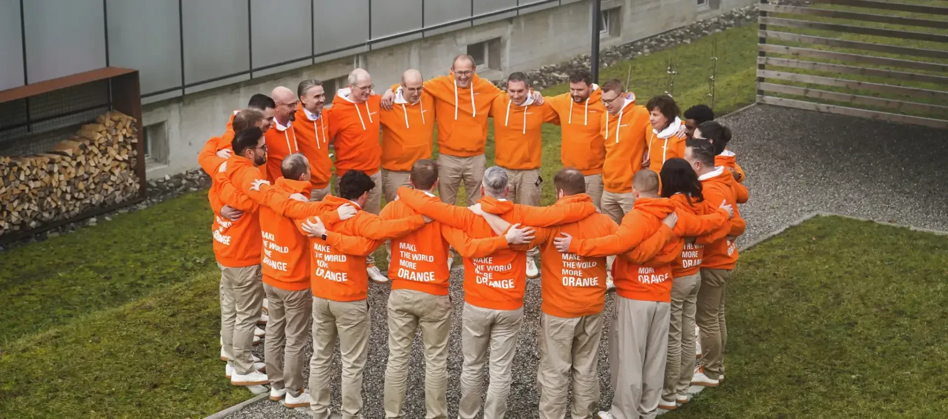 Eine Gruppe von Menschen in orangefarbenen Kapuzenpullis und beigen Hosen steht im Freien in einem Kreis, die Arme umeinander gelegt, auf einer Wiese in der Nähe eines Gebäudes zusammen.