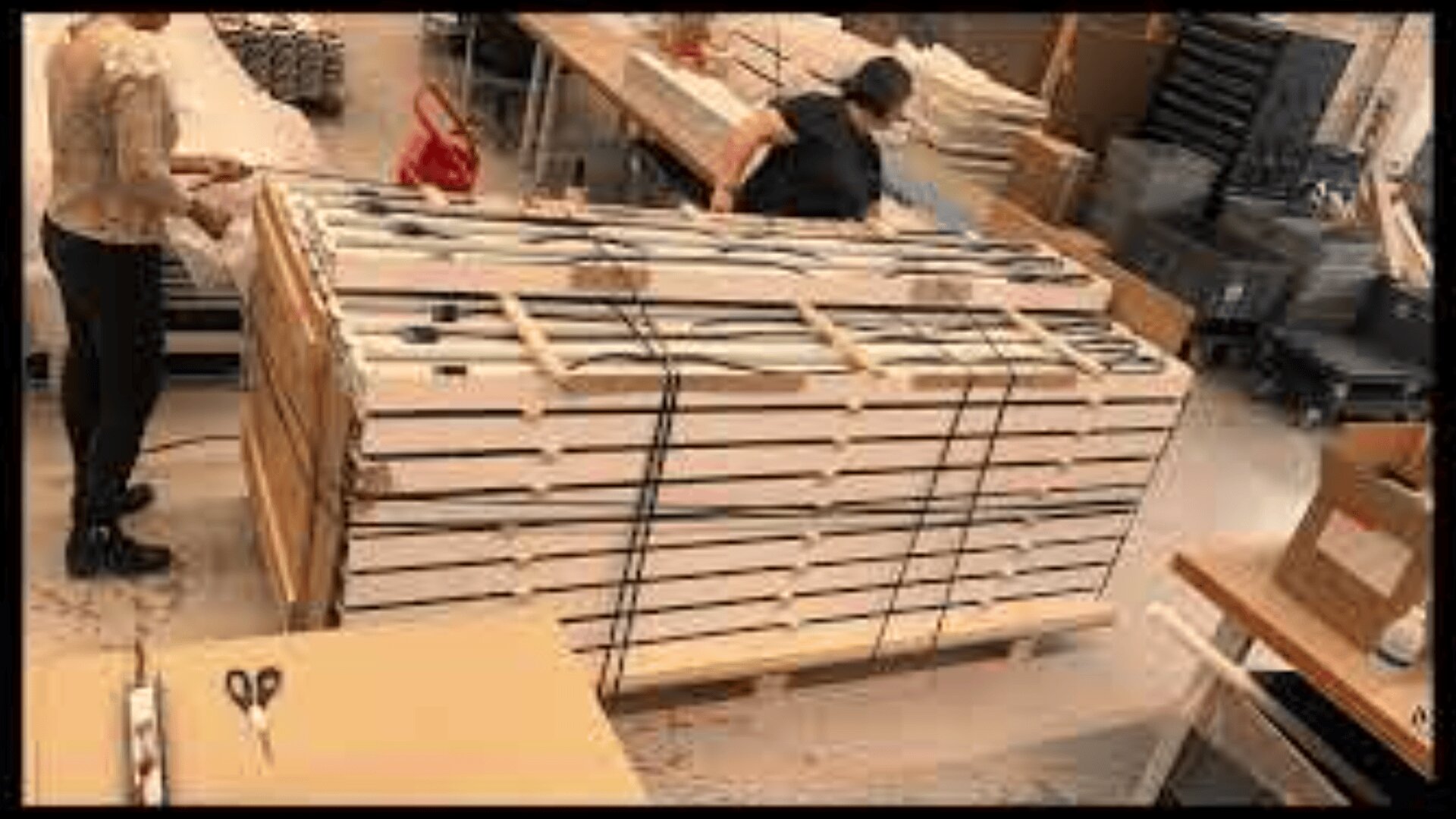 Deux personnes travaillent dans un atelier, emballant de grandes piles d'articles rectangulaires blancs sur une palette en bois, fixée par des sangles noires. Des tables et divers matériaux sont visibles à l'arrière-plan.