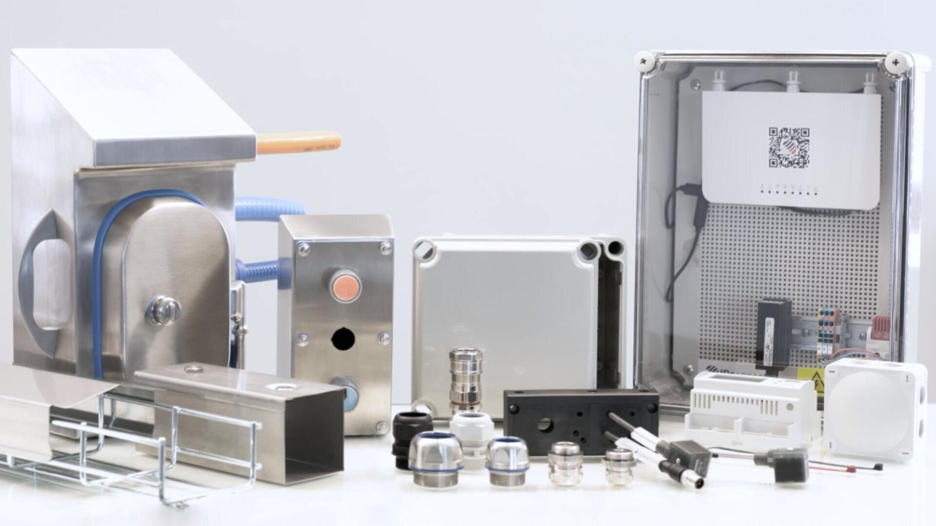 Une variété de boîtiers électriques industriels, de connecteurs, d'interrupteurs, de câbles et d'accessoires de câblage disposés sur une surface blanche, y compris des composants en acier inoxydable et en plastique de différentes formes et tailles.
