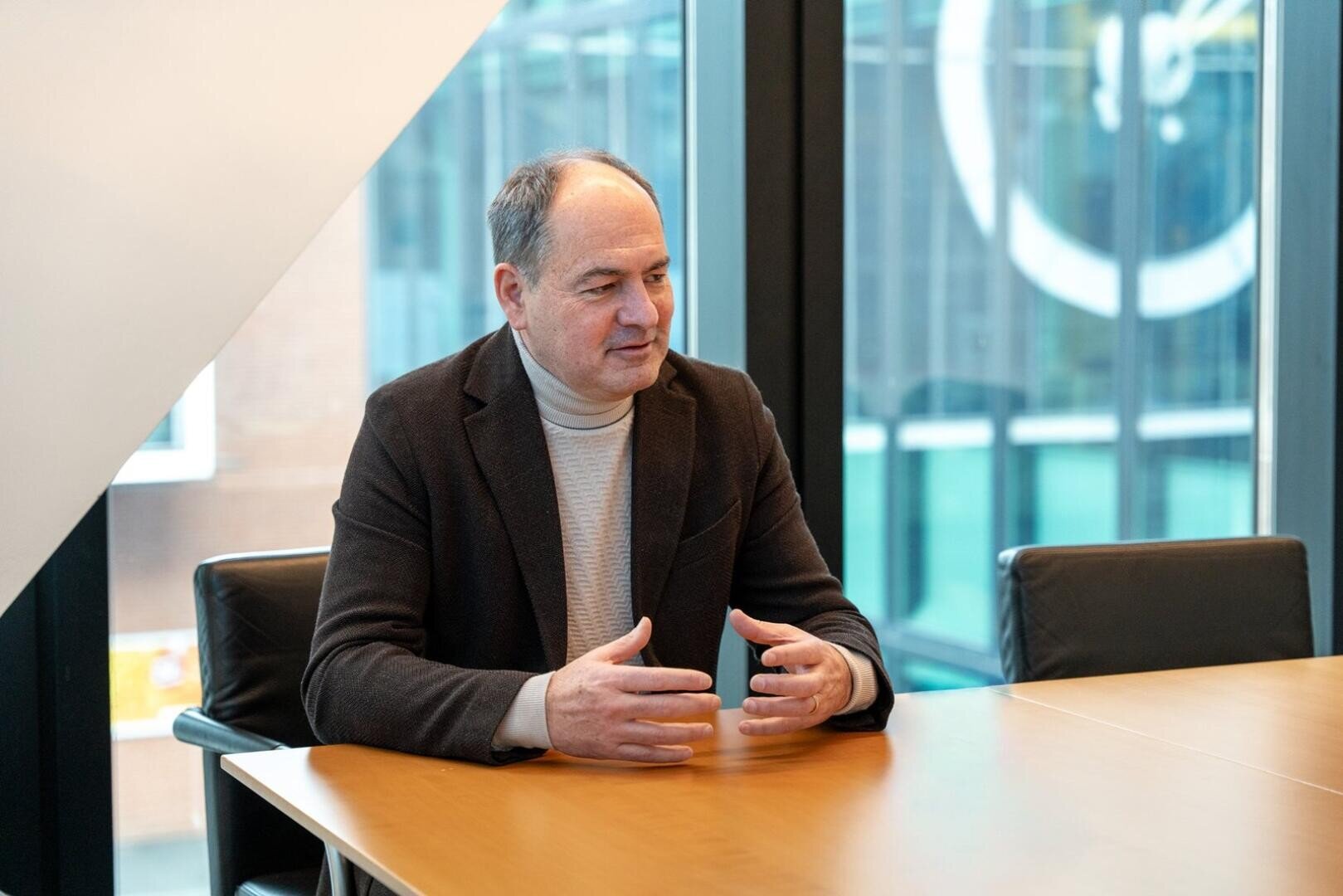Ein Mann in einem braunen Blazer und einem hellen Rollkragenpullover sitzt an einem Konferenztisch und gestikuliert mit seinen Händen, während er in einem modernen Büro mit großen Fenstern im Hintergrund spricht.