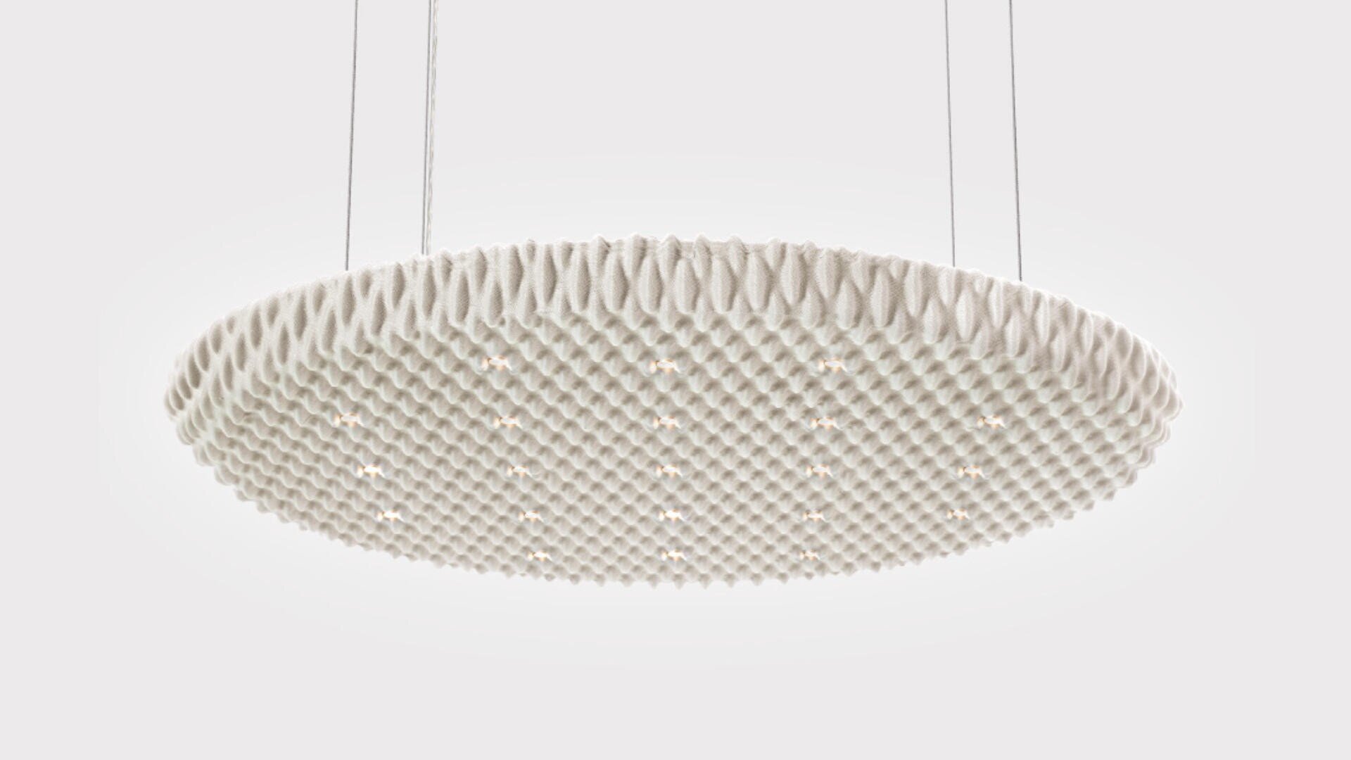 Une grande suspension blanche, ronde et texturée est suspendue au plafond par quatre fils fins. De petites lumières sont réparties uniformément sur la face inférieure et la surface présente un motif géométrique en forme de gaufre.