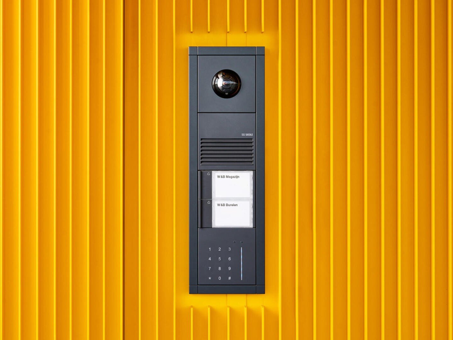 An einer leuchtend gelben, vertikal geriffelten Wand ist eine moderne schwarze Gegensprechanlage, die Vario-Anlage der Firma Siedle, mit Kamera, Lautsprecher, Namensschildern und Zahlen-Tastatur angebracht.