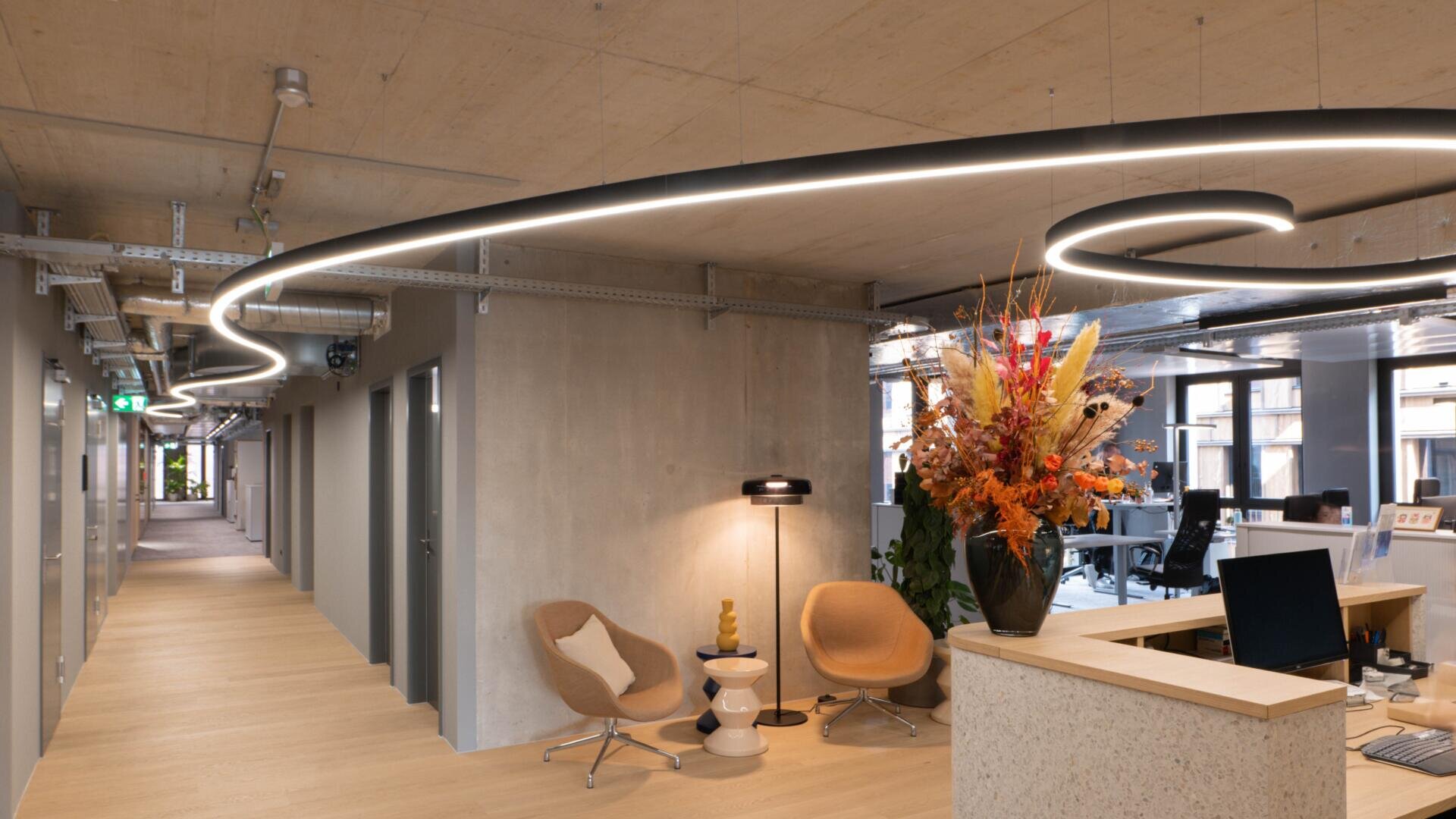Moderne Büroeinrichtung mit Holzböden, Sichtbetonwänden und -decken sowie einer spiralförmigen LED-Leuchte. Zwei hellbraune Stühle und eine Lampe stehen neben einem Empfangstresen, der mit einer großen Vase voller bunter Trockenblumen geschmückt ist.