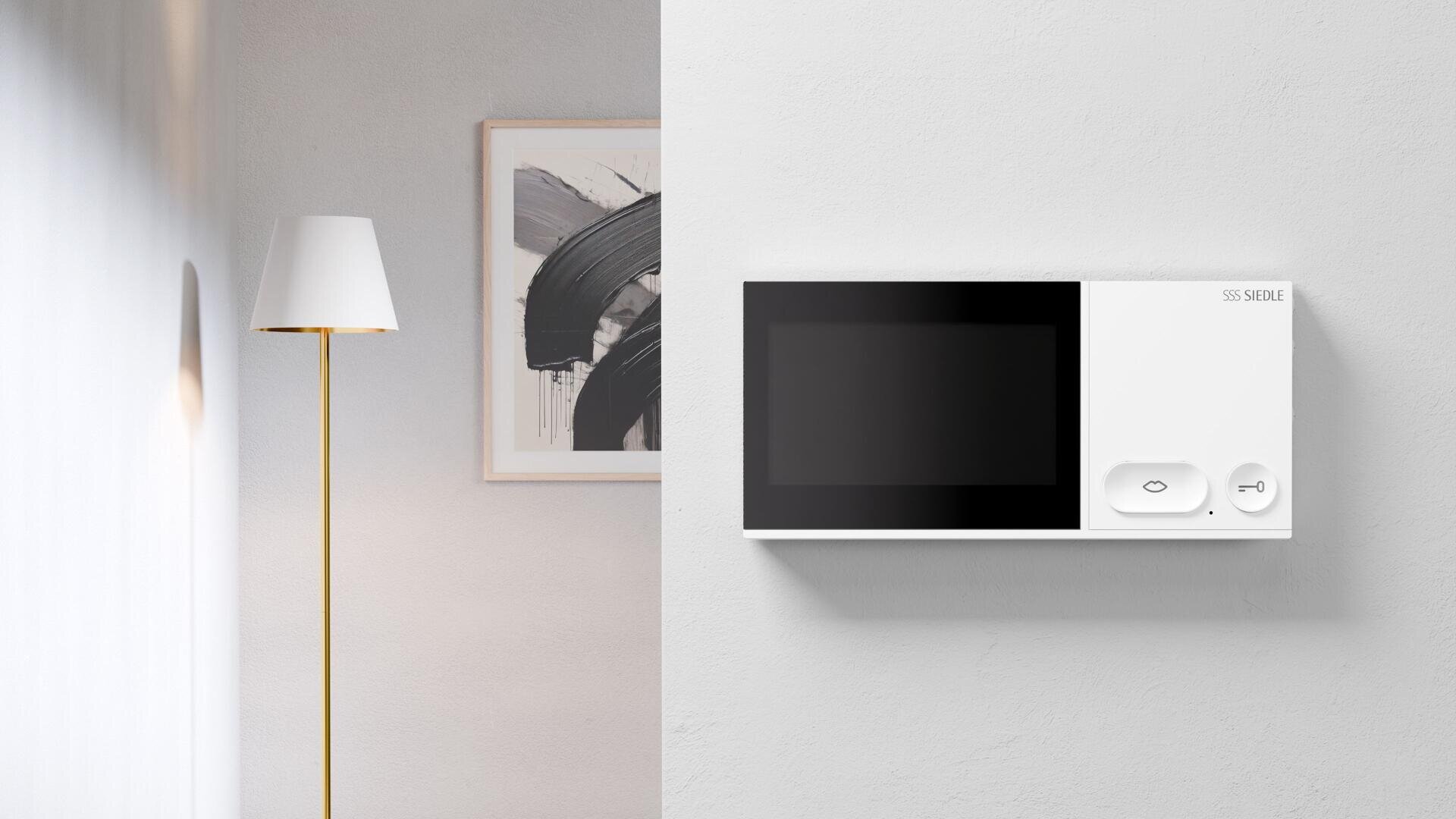 Dans une pièce à la décoration minimaliste, un appareil mural blanc muni d'un écran et de touches se trouve à côté d'un lampadaire doré et d'une œuvre d'art abstraite en noir et blanc.