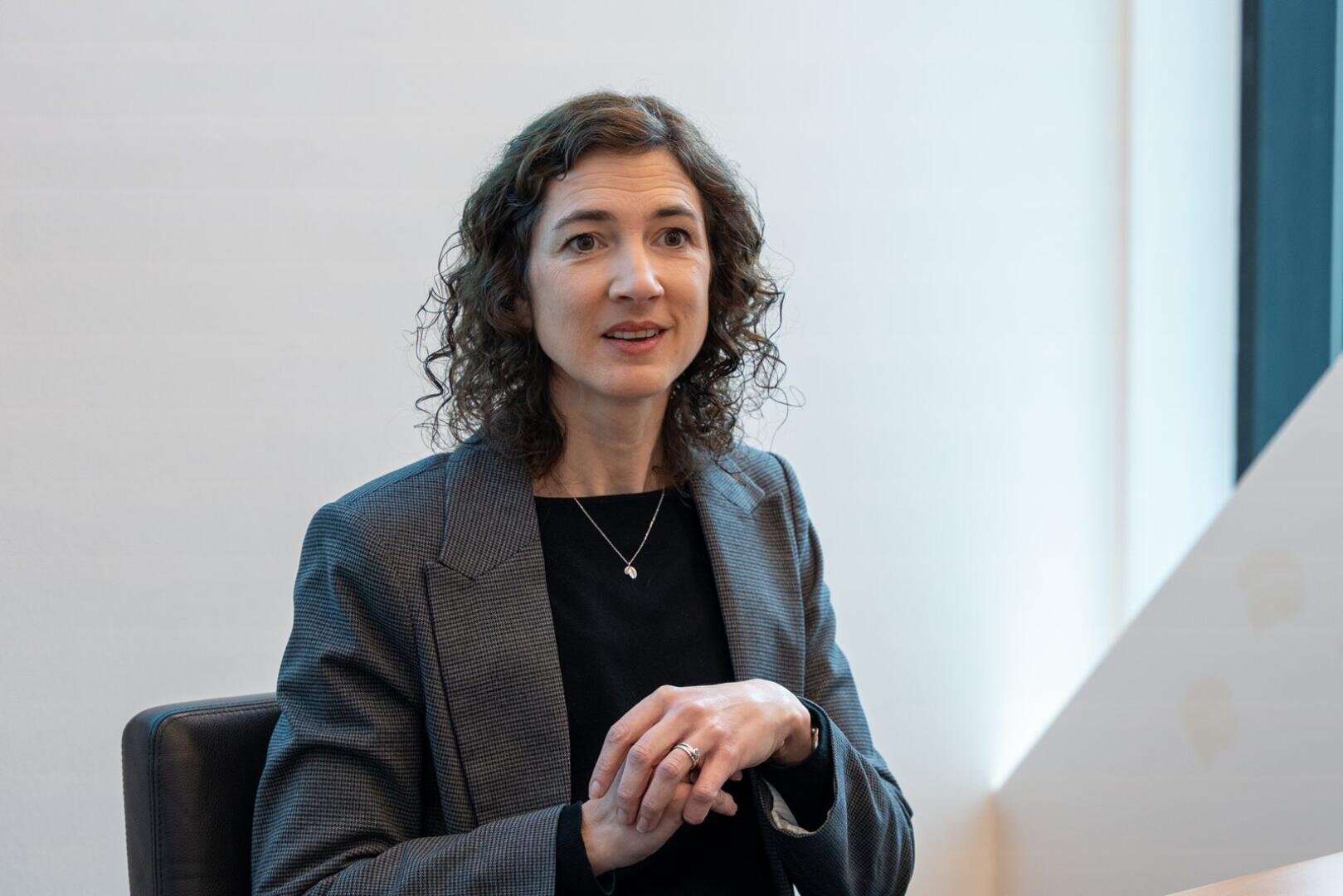 Eine Frau mit lockigem braunem Haar, die einen grauen Blazer und ein schwarzes Oberteil trägt, sitzt an einem Tisch und gestikuliert mit ihren Händen, während sie in einem hellen, modernen Büro spricht.