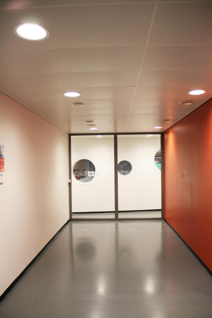 Ein sauberer, moderner Flur mit weißen und orangefarbenen Wänden, grauem Boden und einer Decke mit eingebauten Lichtern, der in Glastüren mit runden Fenstern endet.