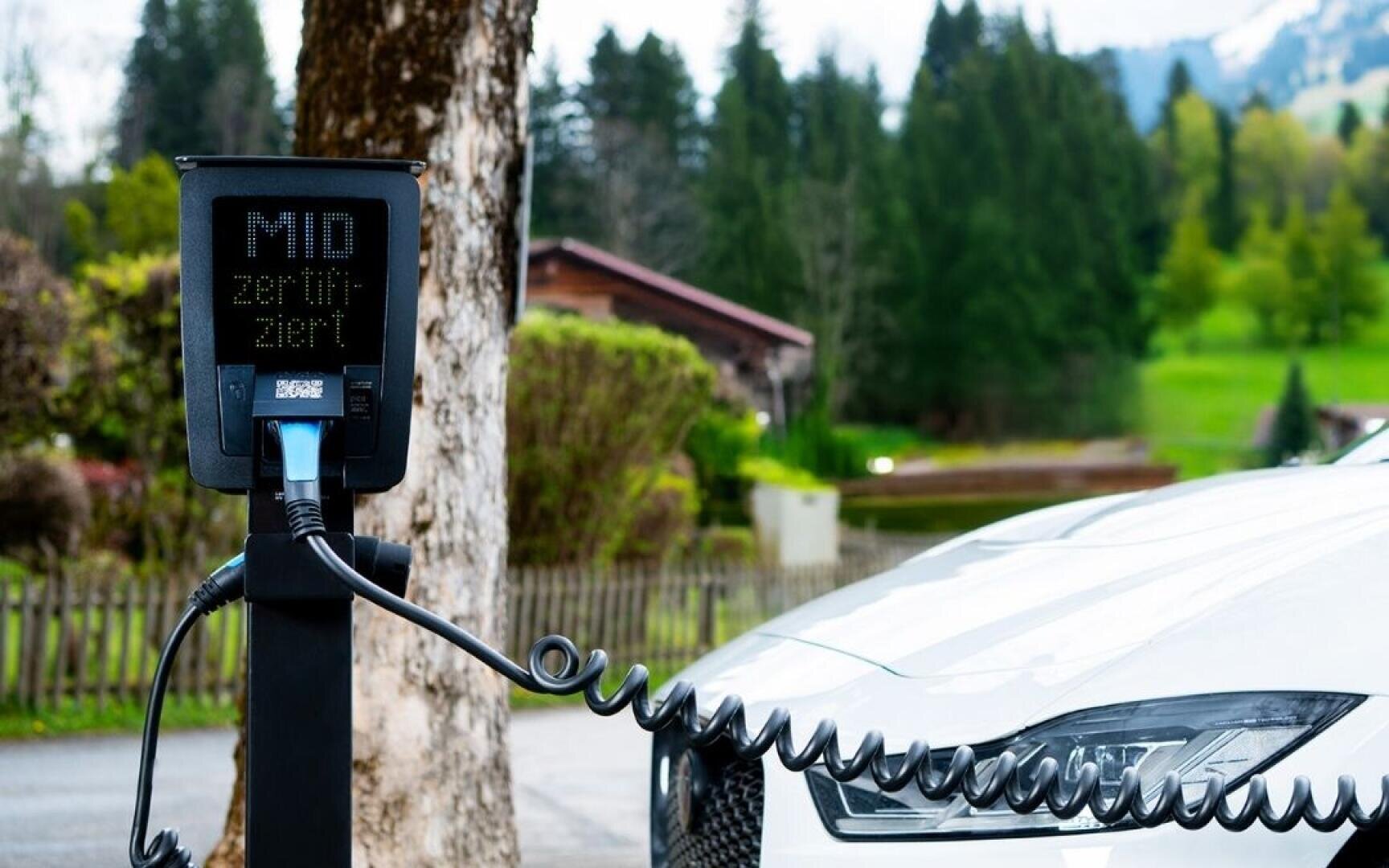 Une voiture électrique est branchée sur une station de recharge en plein air, avec des arbres, de l'herbe et des maisons en arrière-plan. L'écran du chargeur affiche un texte en allemand.
