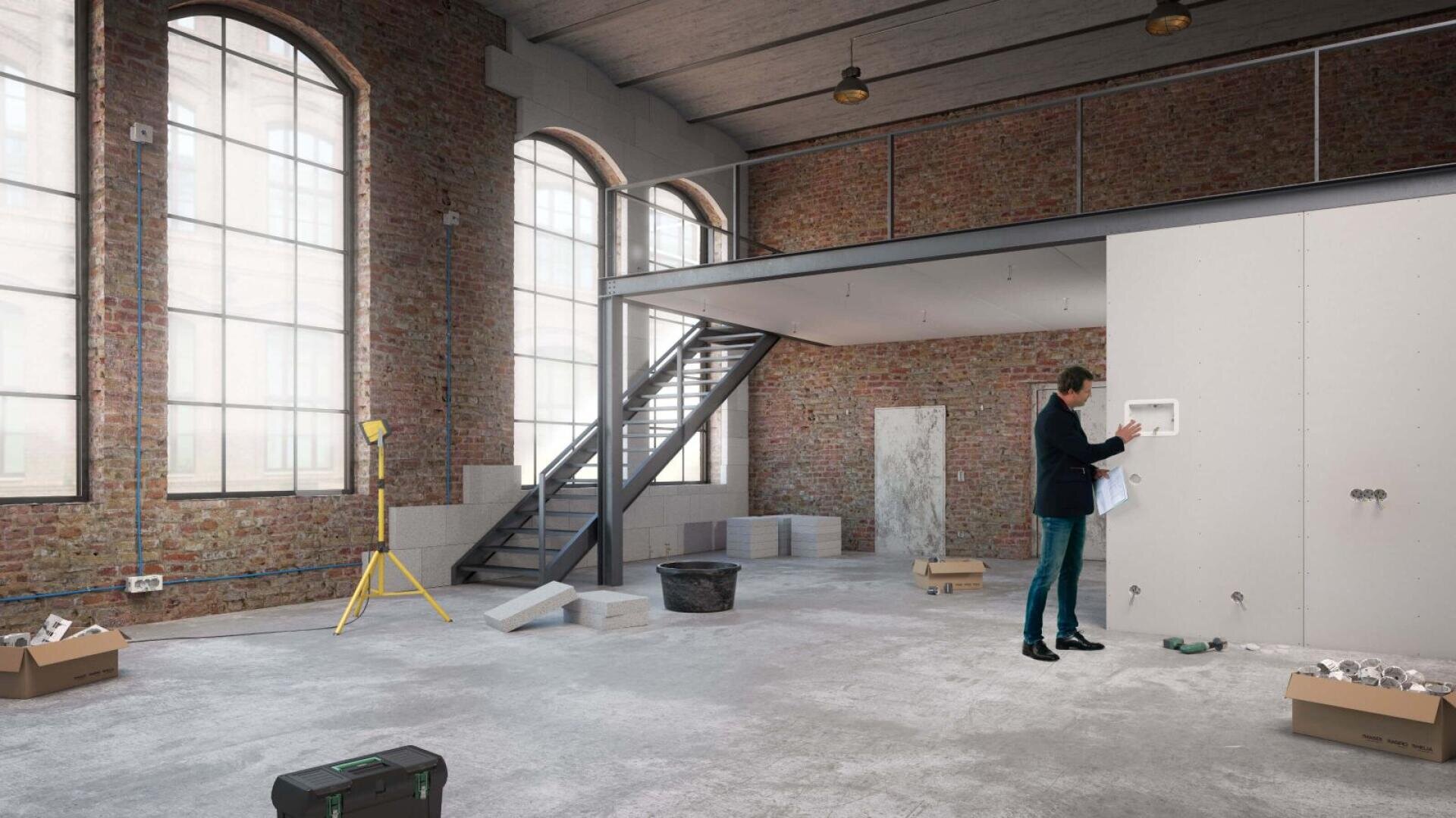 Un homme en costume examine des plans de construction dans une grande pièce industrielle inachevée, avec des murs en briques, de grandes fenêtres cintrées, des matériaux de construction et des outils éparpillés un peu partout.
