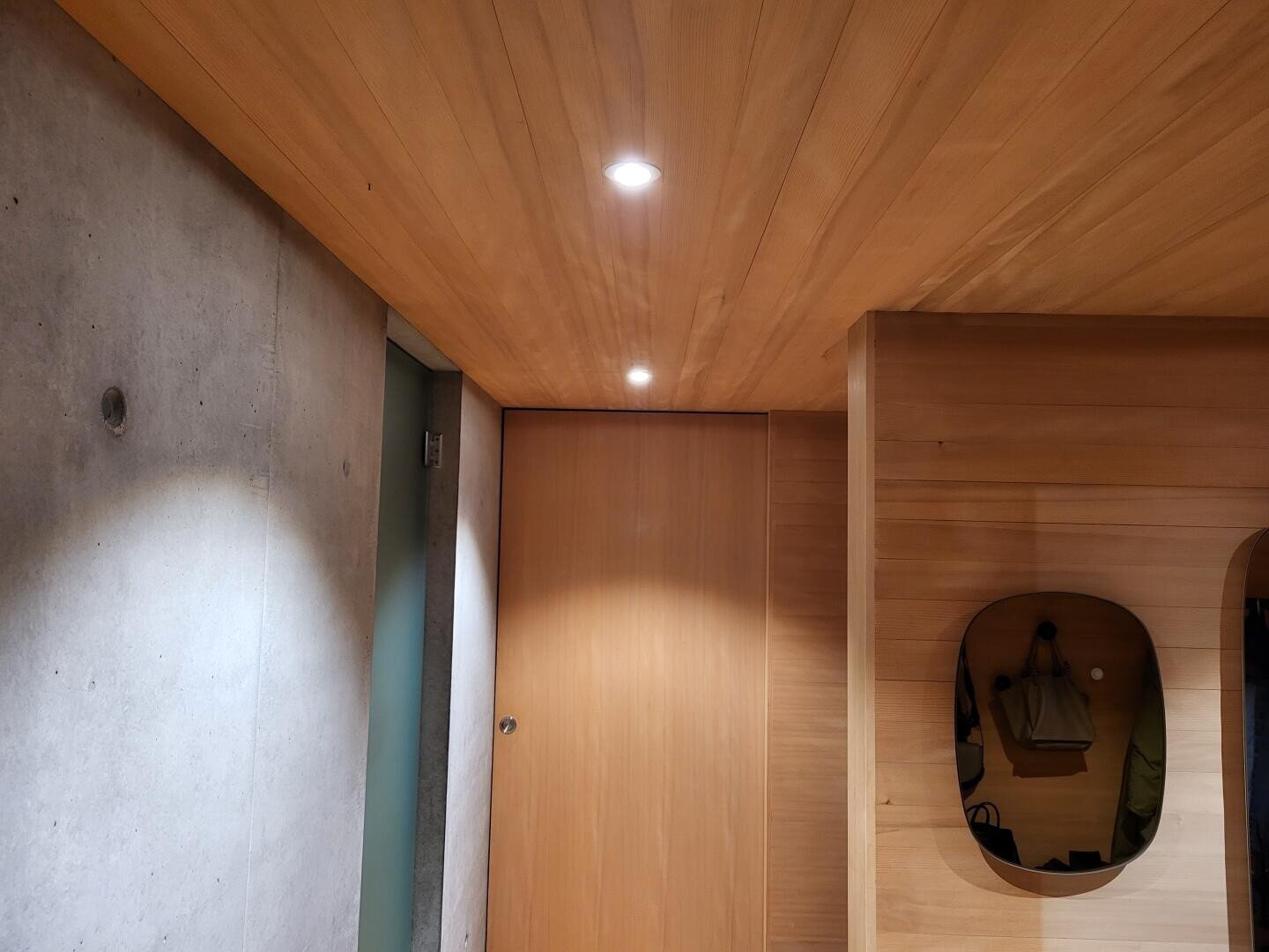 Un intérieur moderne avec un plafond et des murs en bois, des lampes encastrées, une porte fermée, un mur en béton et une petite niche où est suspendu un sac.