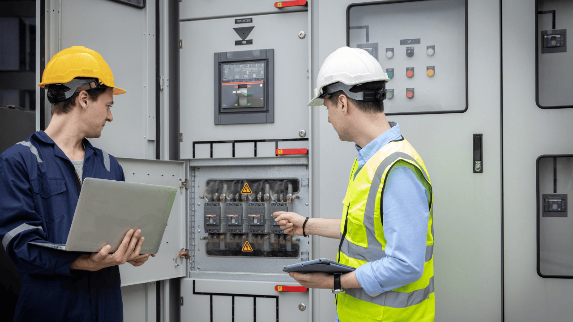 Zwei Ingenieure mit Schutzhelmen und Sicherheitsausrüstung inspizieren elektrische Schalttafeln; einer hält einen Laptop in der Hand, während der andere auf Stromkreisunterbrecher zeigt, beide stehen vor Industrieanlagen mit Warnhinweisen.