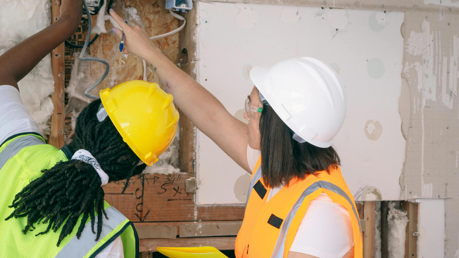 Zwei Bauarbeiter, die Schutzhelme und -westen tragen, untersuchen die Verkabelung in einer zu renovierenden Wand; einer zeigt auf die Drähte, während der andere Werkzeuge oder Geräte hält.