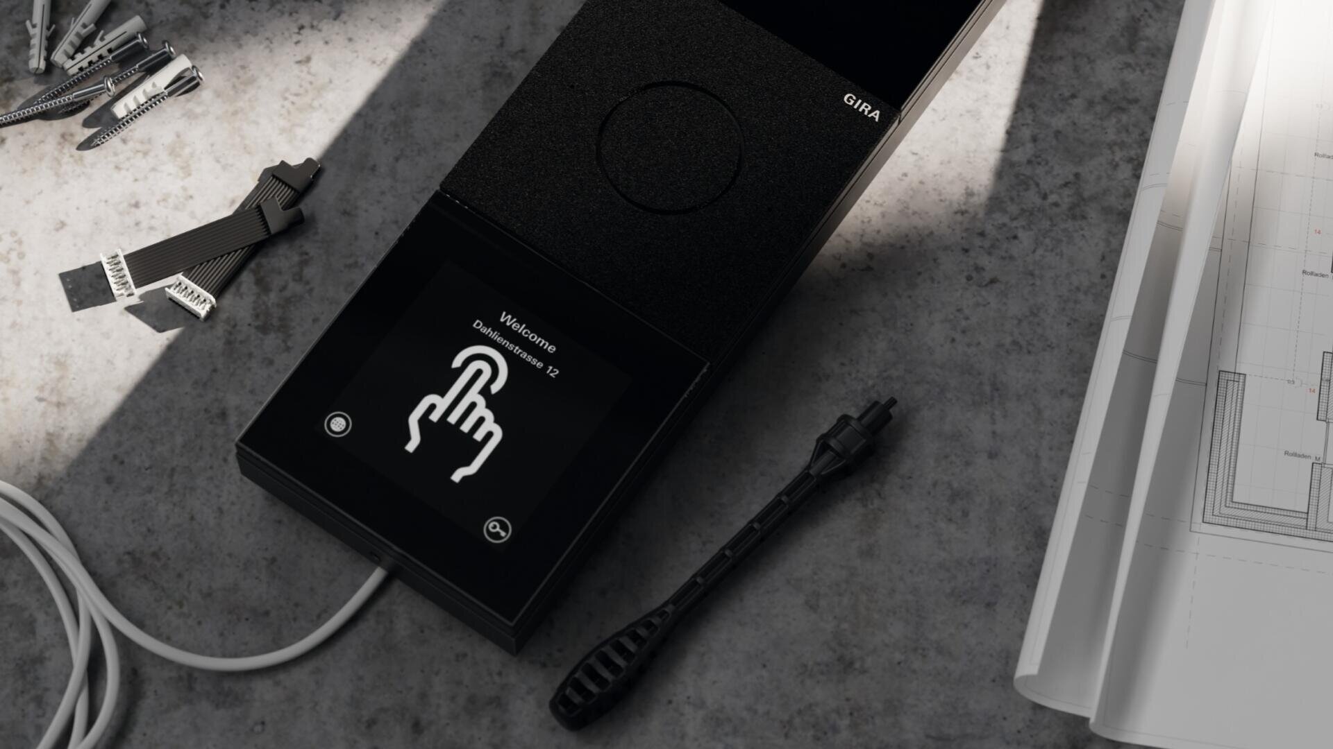 Ein schwarzes Touchscreen-Gerät auf einer grauen Oberfläche zeigt eine Willkommensmeldung und ein Handsymbol an, das auf eine Schaltfläche drückt. In der Nähe liegen elektronische Bauteile und Kabel verstreut, ebenso wie eine Ecke einer technischen Zeichnung.