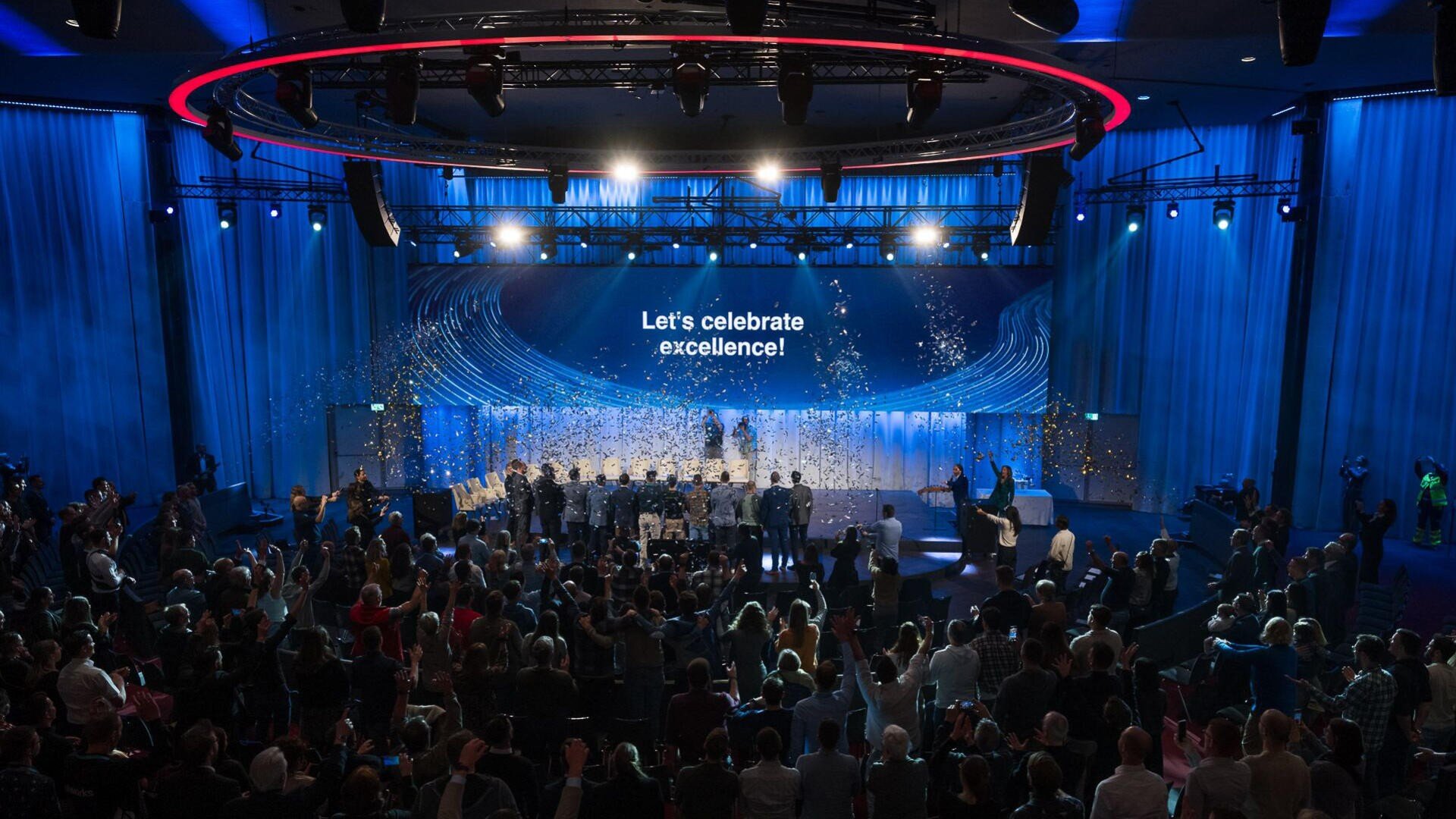 Ein großes Publikum verfolgt eine Bühnenveranstaltung in einem geräumigen, modernen Auditorium. Auf der Leinwand wird "Lets celebrate excellence!" angezeigt, während Konfetti fällt und die Menschen auf der Bühne unter hellen Lichtern gefeiert werden.