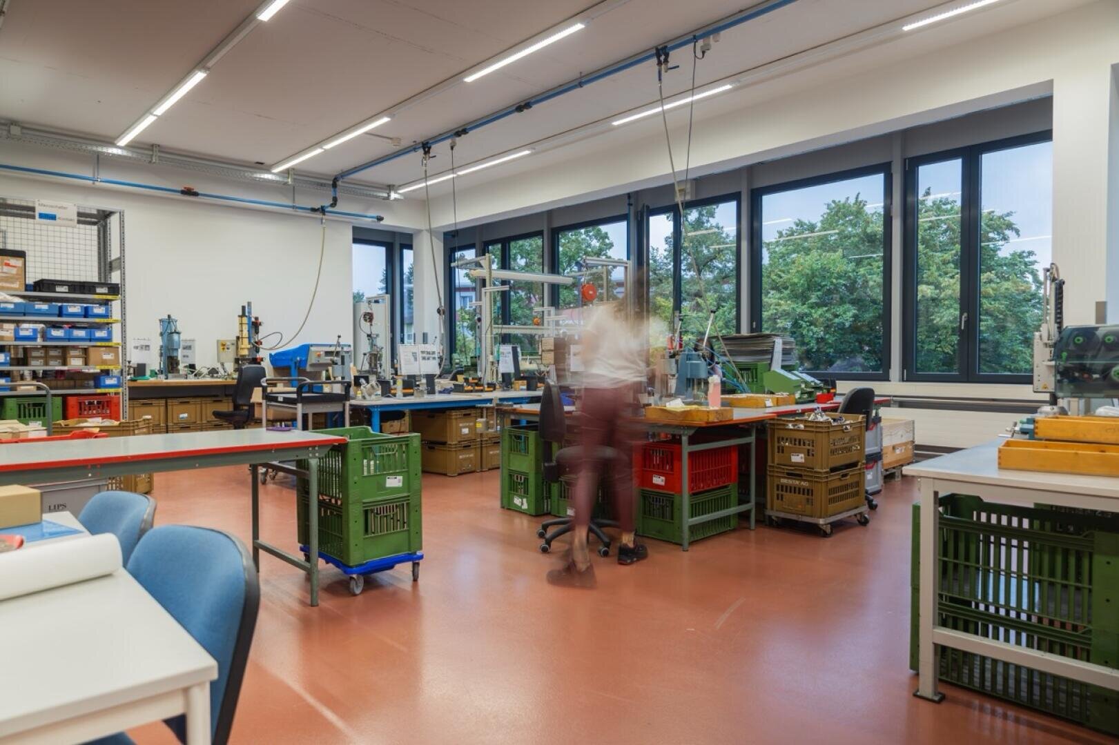 Un atelier spacieux et bien éclairé, équipé de diverses machines, d'outils et d'établis. Des caisses vertes et orange sont empilées un peu partout. De grandes fenêtres donnent sur des arbres à l'extérieur. On aperçoit une personne qui marche, légèrement floue à cause du mouvement.