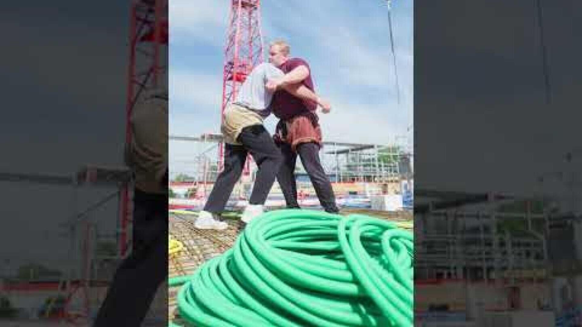 Zwei Personen ringen auf einer Baustelle in der Nähe eines großen grünen Schlauchs, im Hintergrund sind unter blauem Himmel Kräne und Gerüste zu sehen.