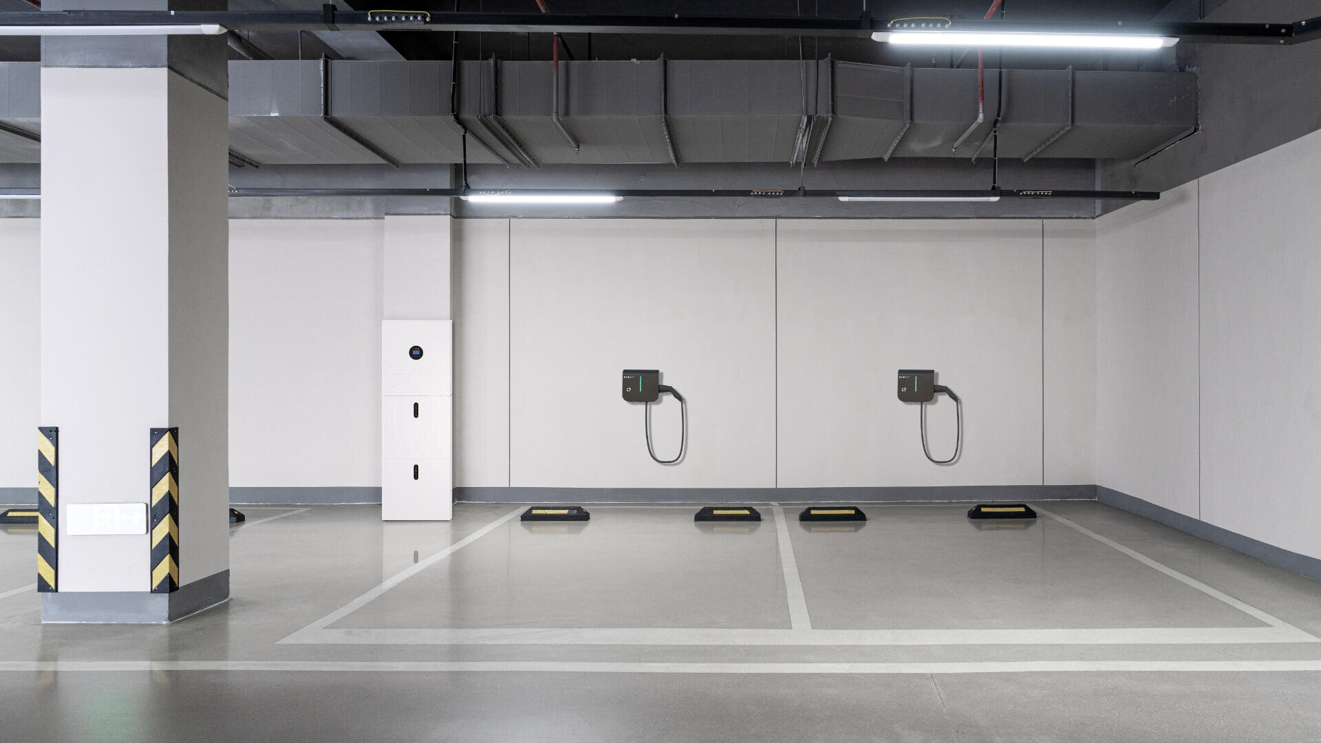 Ein sauberer überdachter Parkplatz mit drei leeren Parkplätzen, die jeweils mit einer an der Wand montierten Ladestation für Elektrofahrzeuge ausgestattet sind. Ein hoher weißer Schrank steht links neben einer Säule.