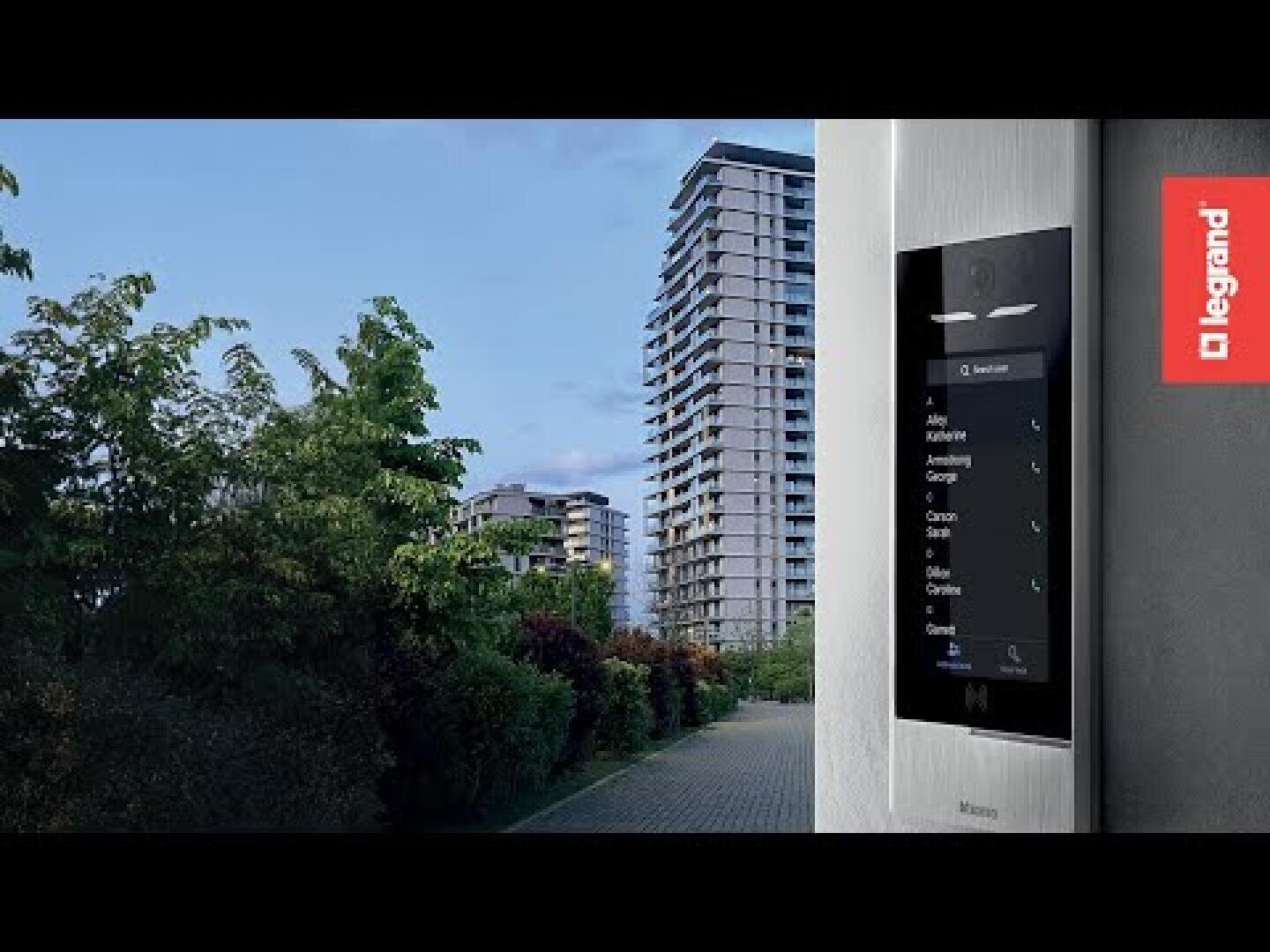 Ein modernes Mehrfamilienhaus unter blauem Himmel mit einer intelligenten Videosprechanlage, die an einer Wand im Vordergrund montiert ist; das Legrand-Logo erscheint auf der rechten Seite des Bildes.