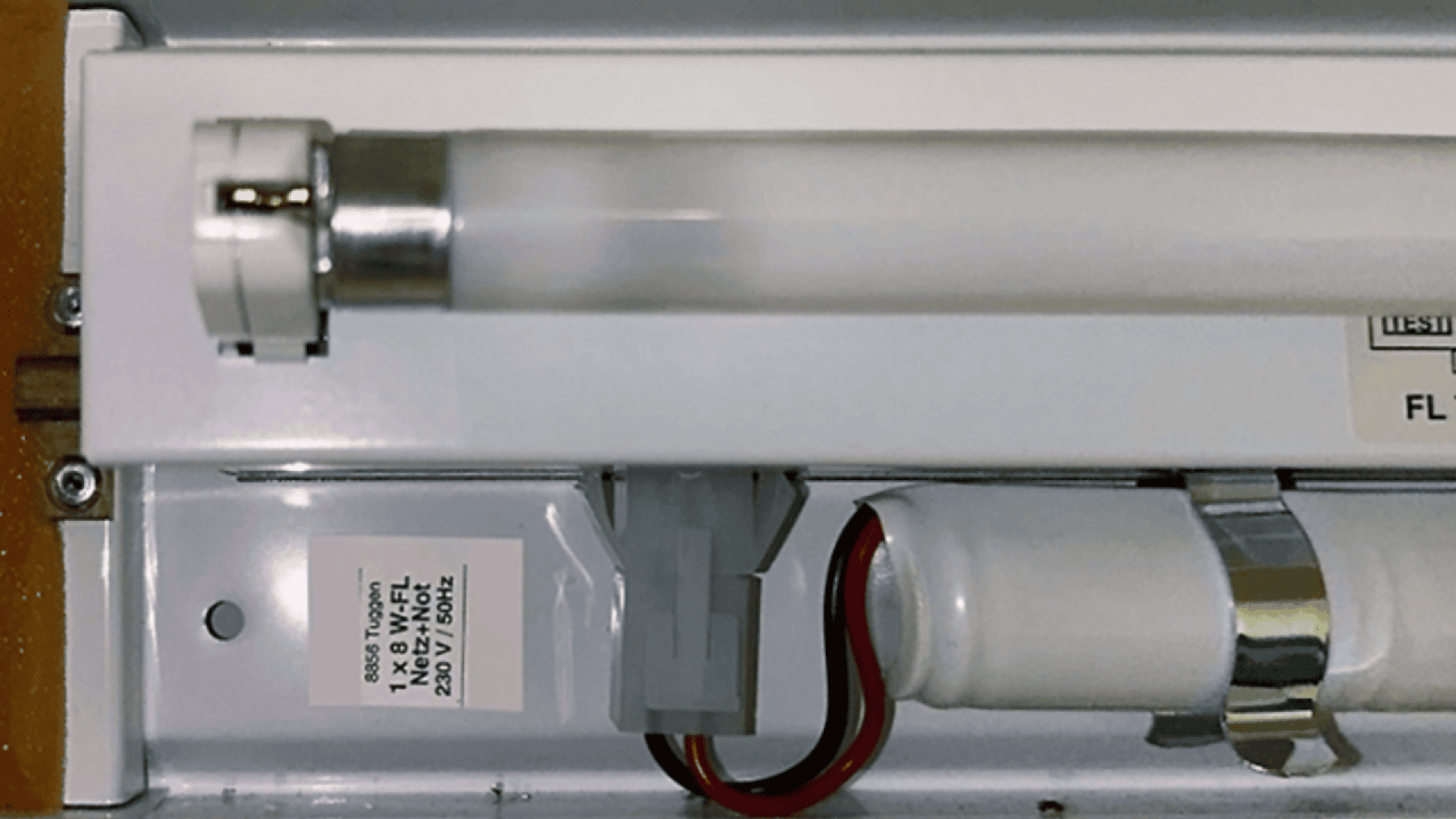 Gros plan d'un luminaire fluorescent montrant le tube, le câblage et le connecteur électrique. Une étiquette contenant des informations sur la tension et le câblage est visible près de la lampe.