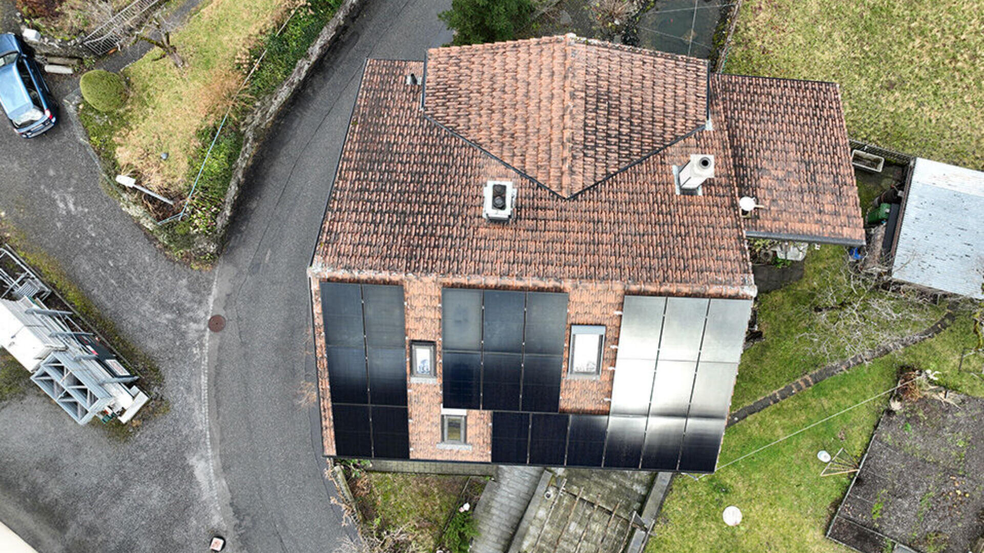 Luftaufnahme eines Hauses mit Ziegeldach und mehreren auf einer Seite installierten Sonnenkollektoren; das Haus ist von einer Straße und Grünflächen umgeben.