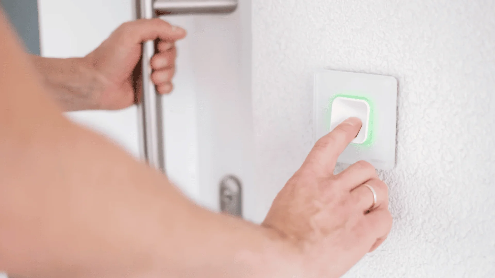 Eine Person drückt eine weiße Türklingel mit grünem Licht an einer weißen Wand, während sie mit der anderen Hand einen Türgriff festhält.