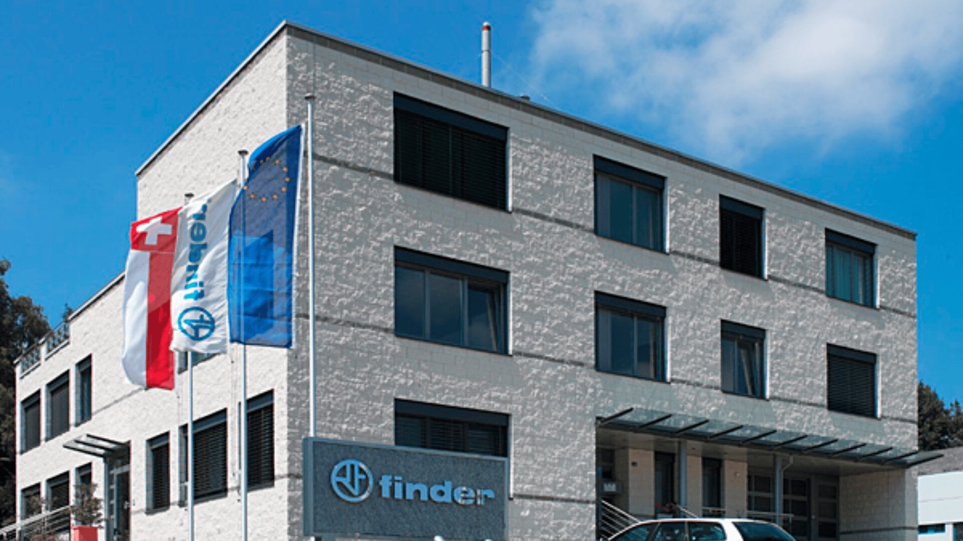 Ein modernes weißes Bürogebäude mit drei Stockwerken, großen Fenstern und einem Finderschild über dem Eingang. Vor dem Gebäude wehen drei Flaggen, darunter die Flagge der Schweiz und der Europäischen Union, unter blauem Himmel.