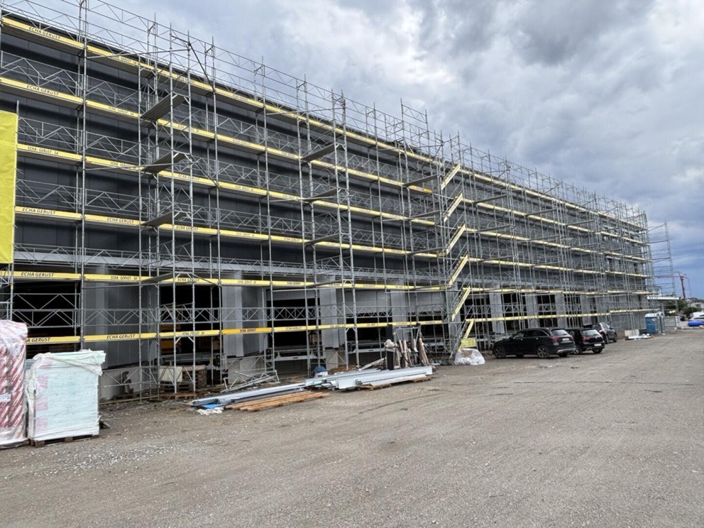 Ein großes Gebäude, das sich im Bau befindet, ist mit Gerüsten umgeben. In der Nähe liegen Baumaterialien auf dem Boden verstreut, und unter einem bewölkten Himmel stehen mehrere Autos entlang der Baustelle.