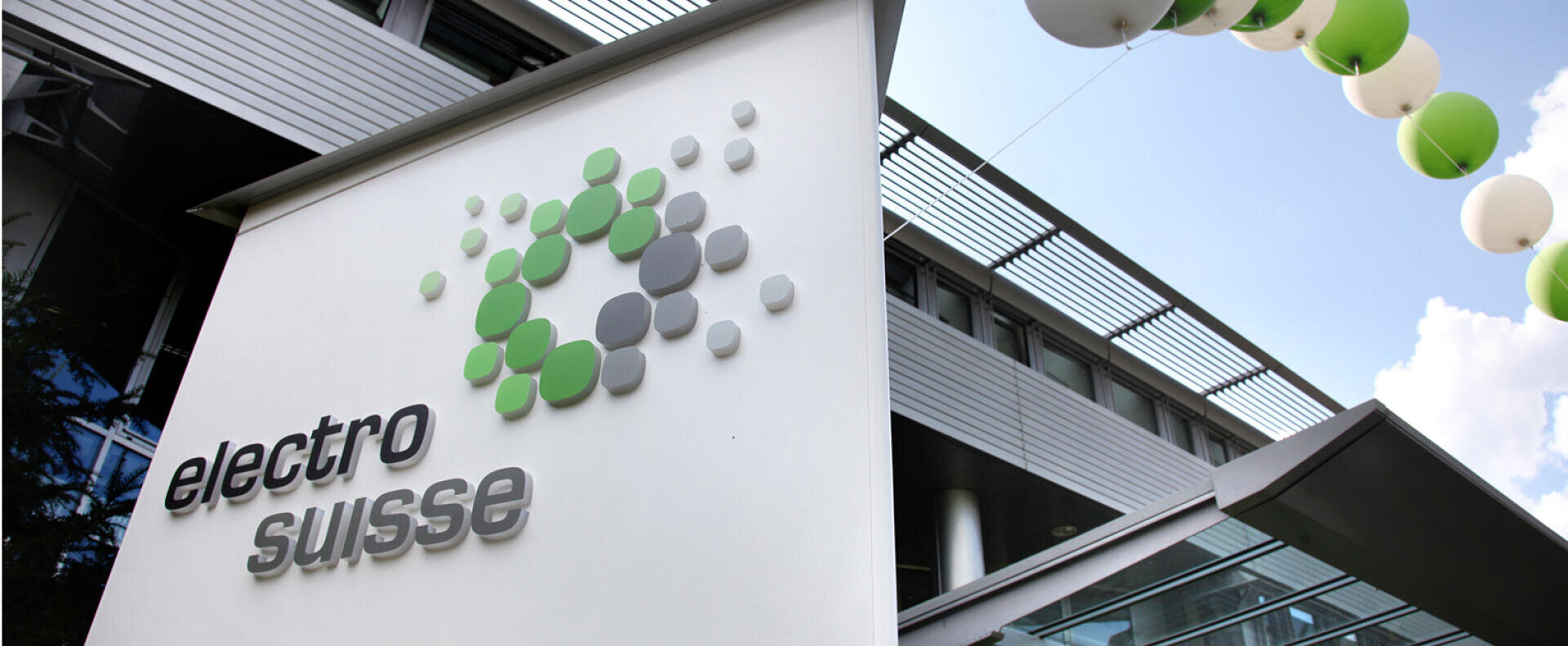 Ein modernes Gebäude mit einem Schild mit der Aufschrift „electro suisse“ und einem Logo aus grünen, grauen und weißen Punkten. Darüber hängen weiße und grüne Luftballons, und eine Glasdachkonstruktion ist zu sehen. Der Himmel ist teilweise bewölkt.